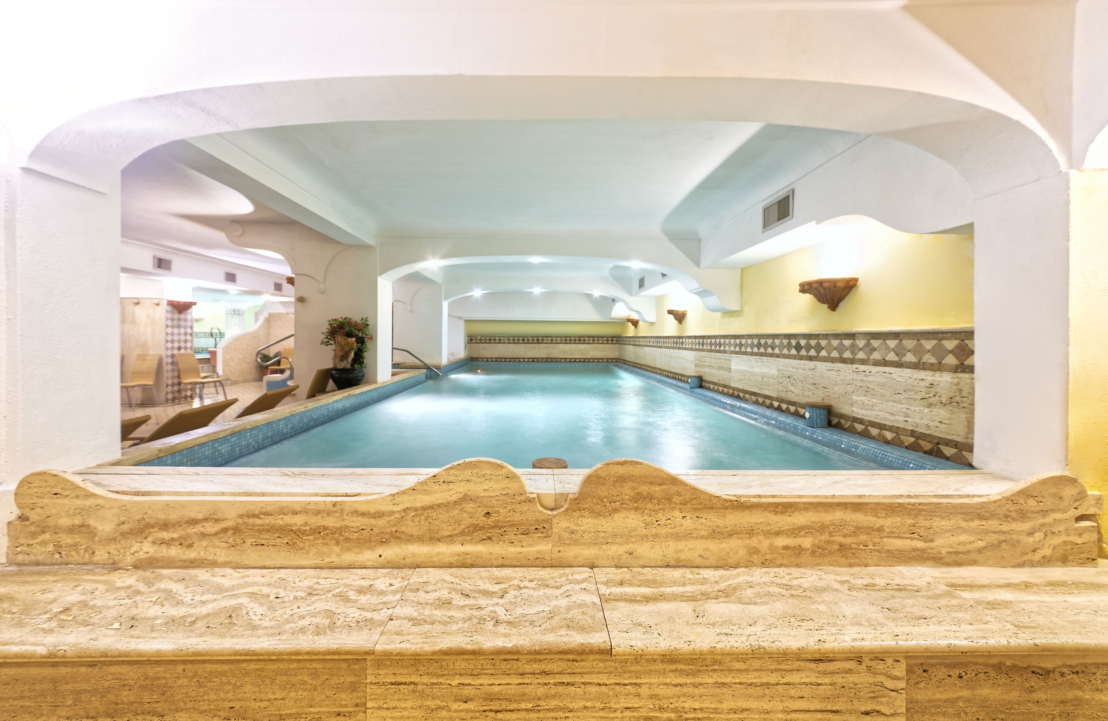 An indoor swimming pool with a stone border, a tiled wall, and a ceiling with recessed lighting.