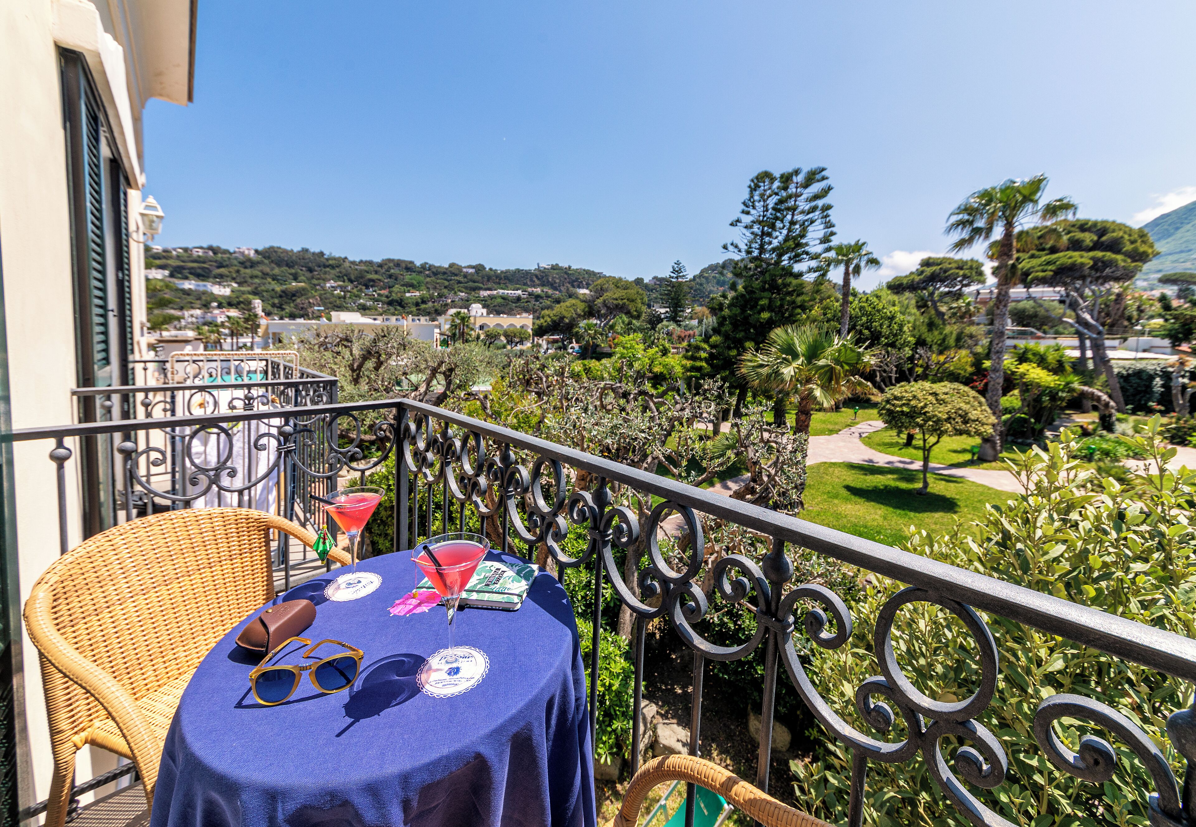 Un balcon avec une table dressée pour deux personnes, offrant une vue sur un jardin luxuriant et des bâtiments situés au loin.
