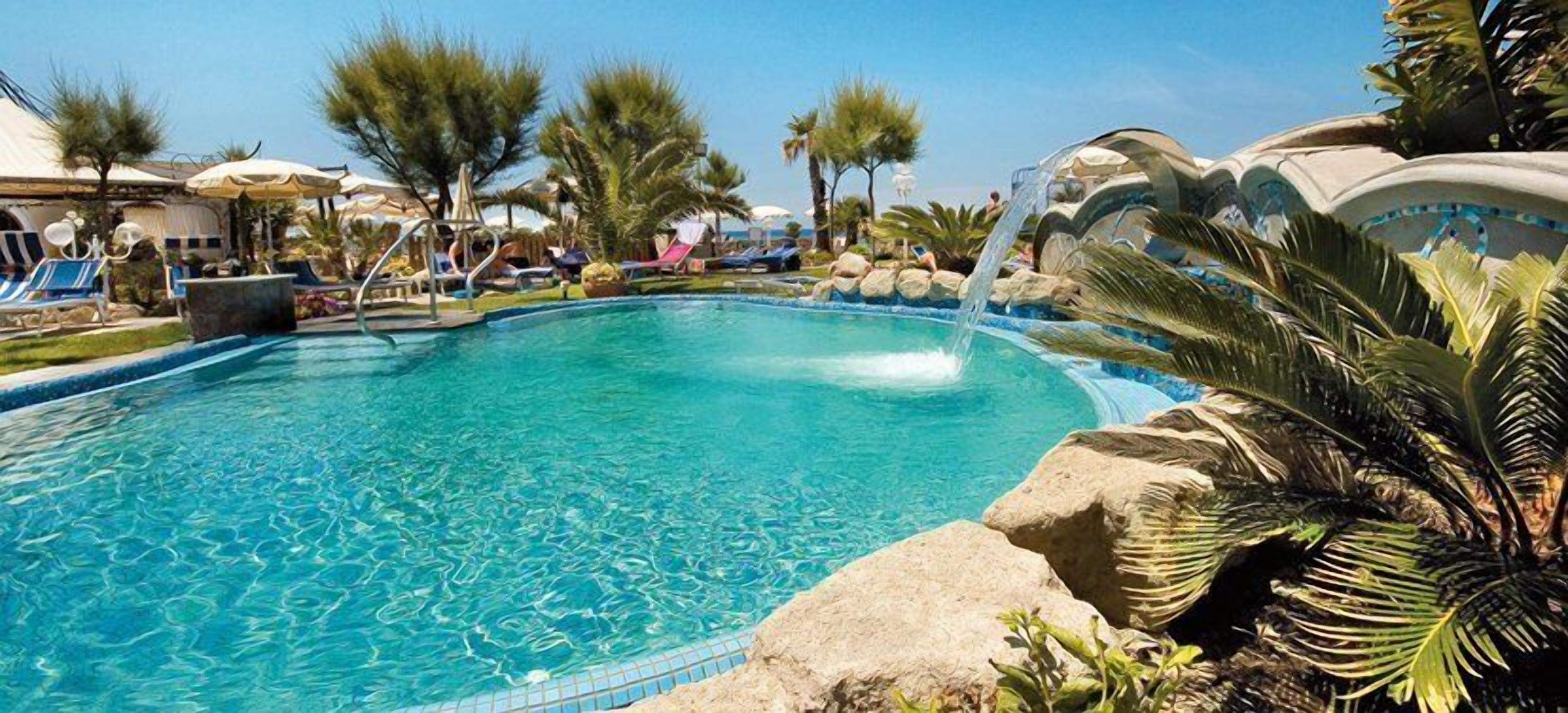 Una piscina cristallina blu con una cascata, circondata da lettini prendisole e palme.