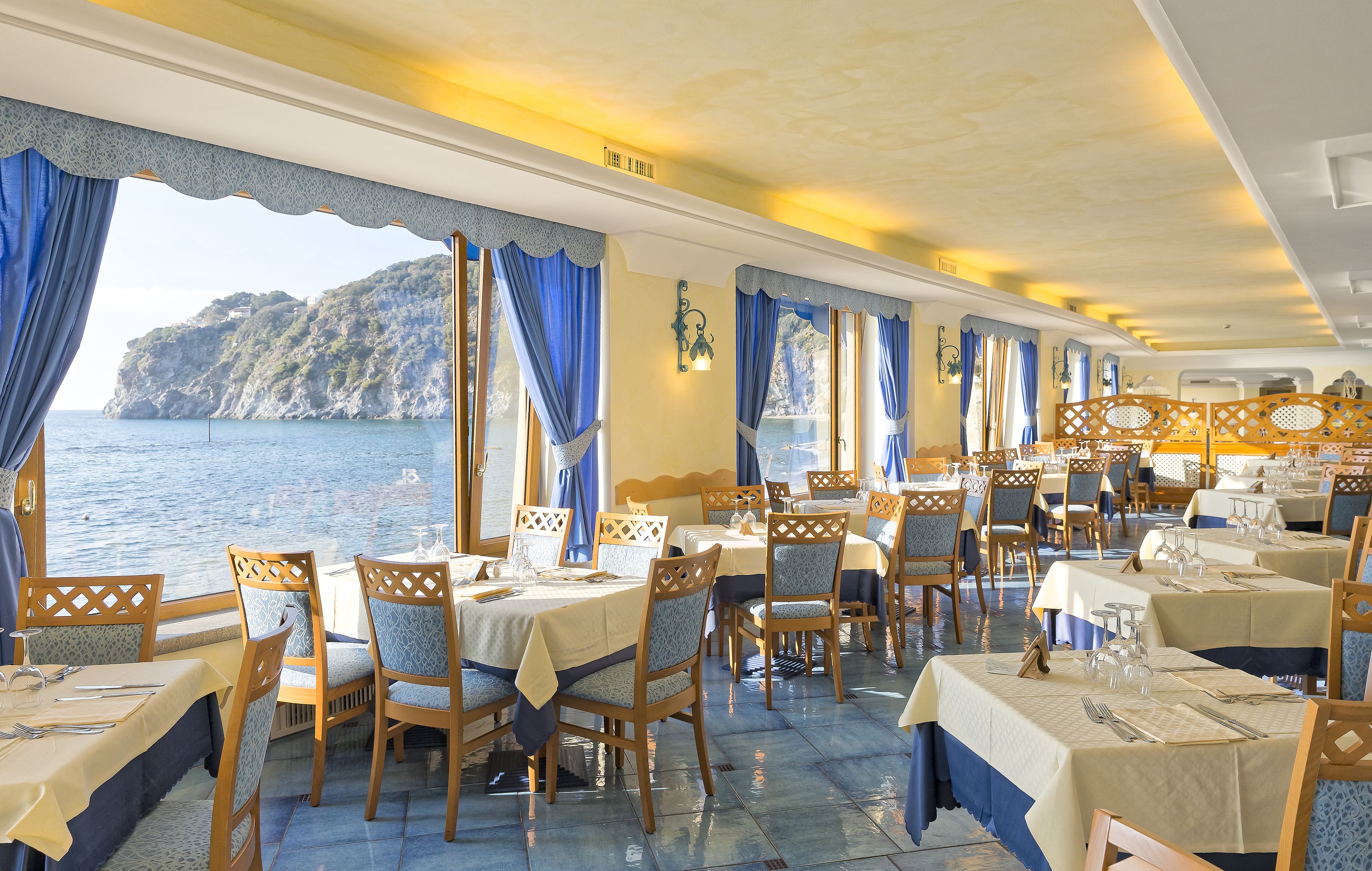 A dining area with tables and chairs, large windows with blue curtains, and a view of the sea and a mountain.