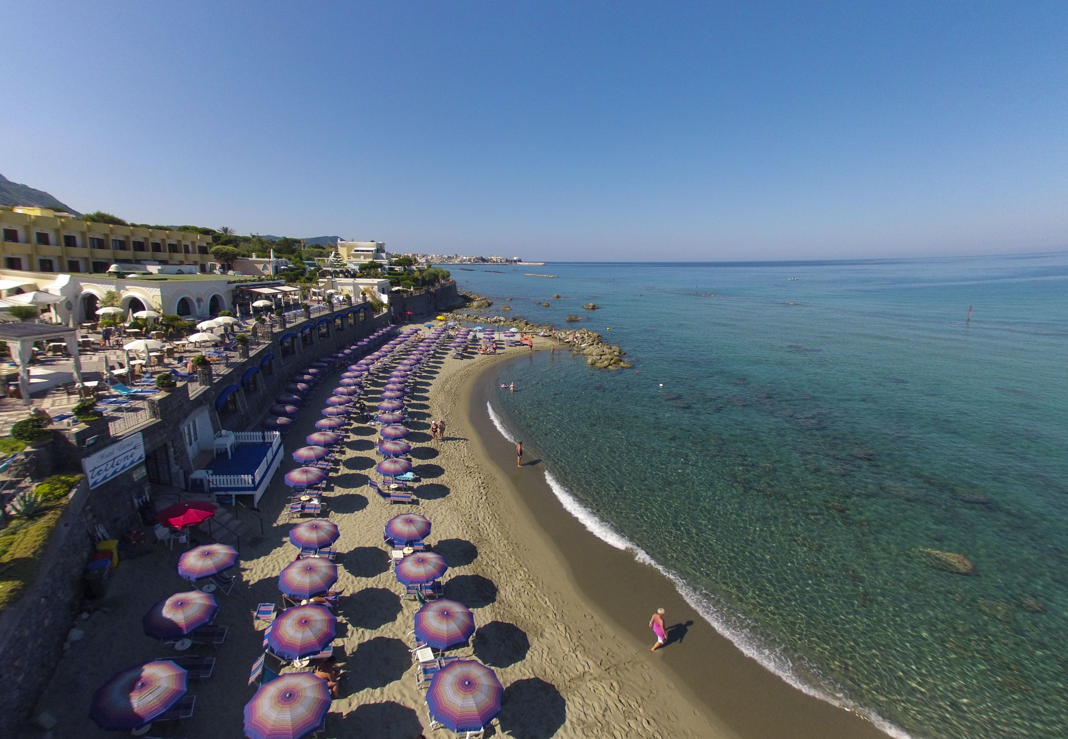 Una playa con hileras de sombrillas y sillas, un paseo marítimo con edificios, y agua azul cristalina.