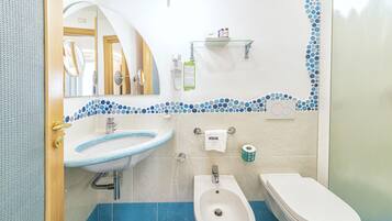 A bathroom with a sink, bidet, and toilet, a mirror, and a shelf with toiletries.