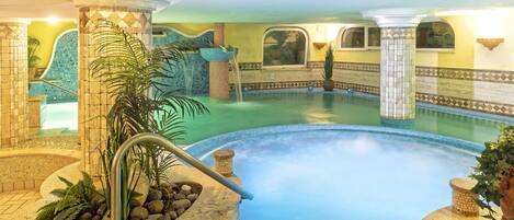 A large indoor swimming pool with a tiled edge, surrounded by stone pillars and plants.