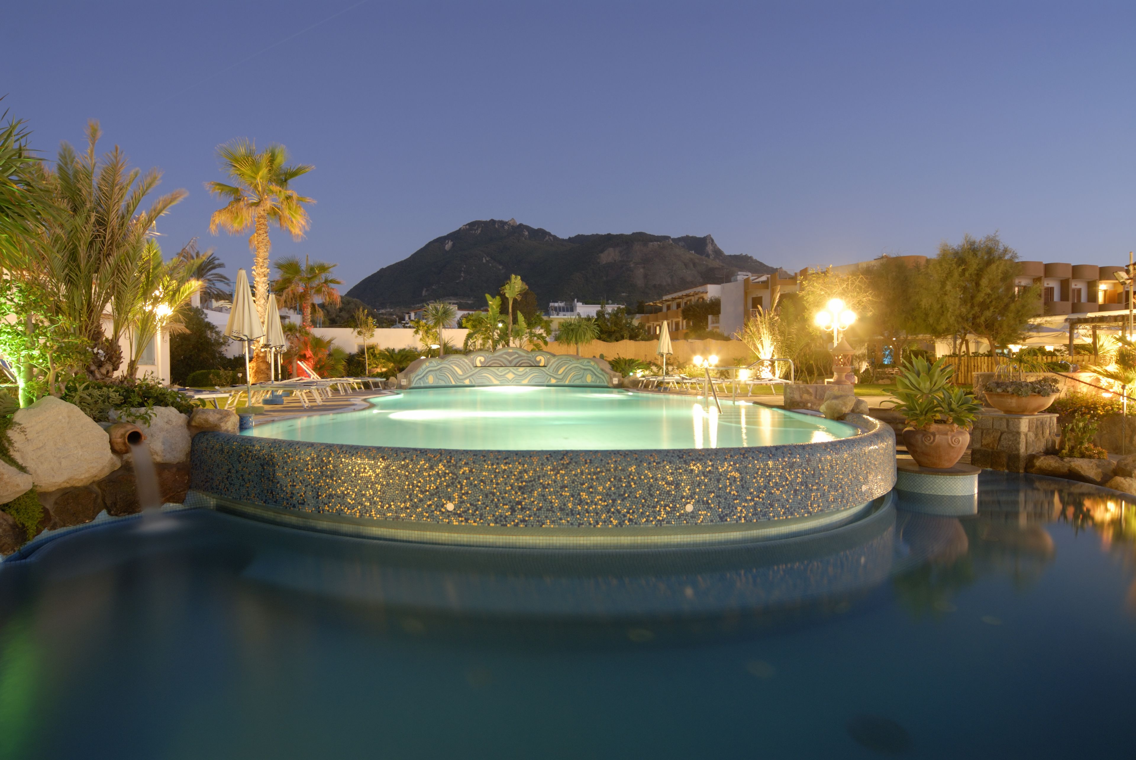 A pool with a pebble edge, surrounded by palm trees and potted plants, with a mountain in the background.