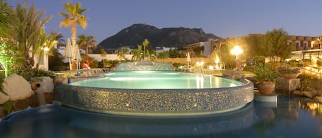 A pool with a pebble edge, surrounded by palm trees and potted plants, with a mountain in the background.