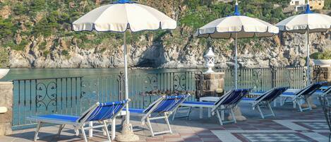 A coastal terrace with sun loungers and umbrellas, a patterned floor, and a rocky hillside in the background.