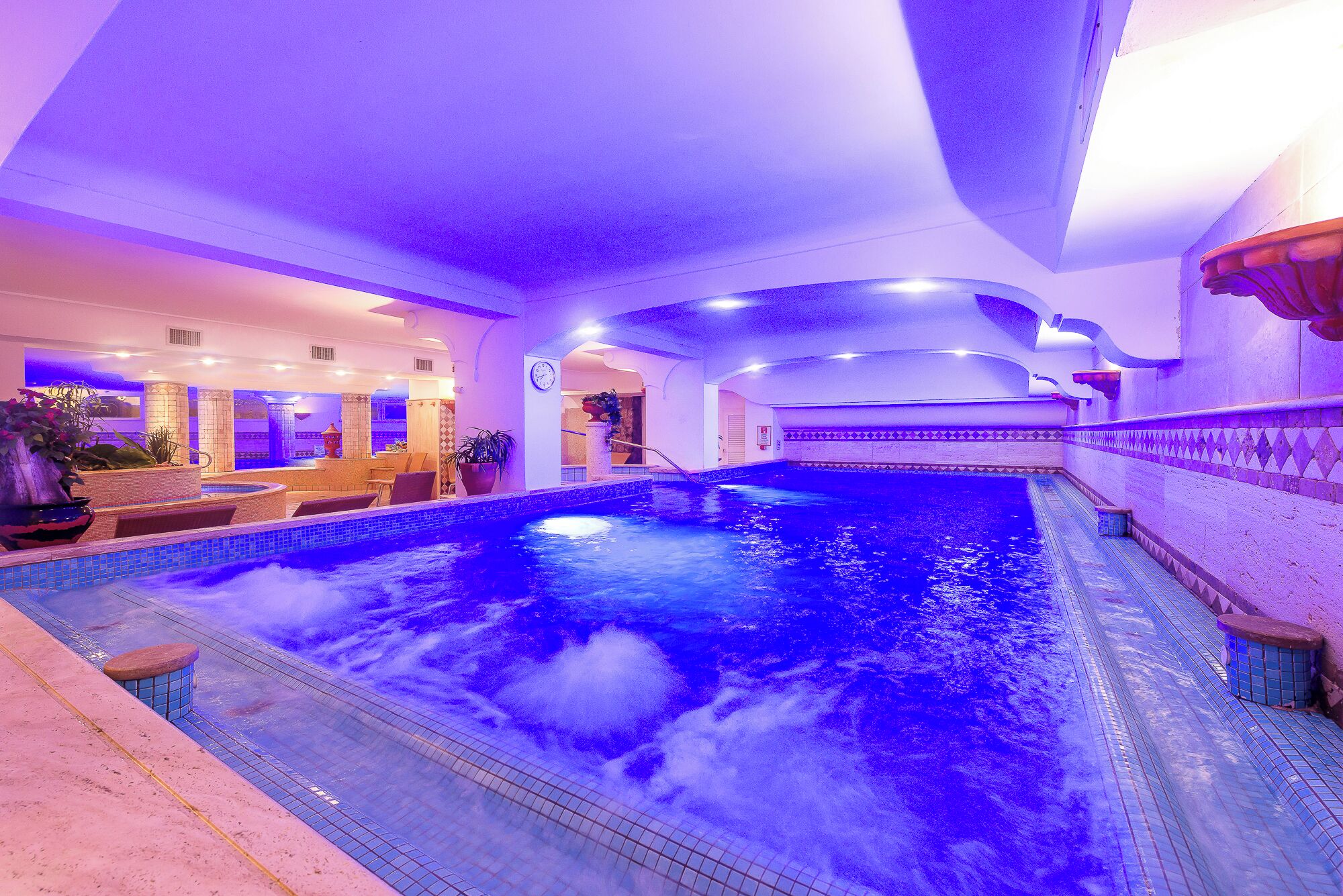 An indoor swimming pool with a blue water feature, surrounded by a tiled area with seating and a small waterfall.