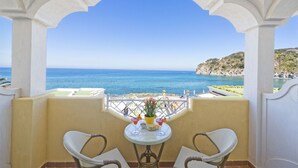 A balcony with two wicker chairs, a small round table with a vase and drinks, and a view of the sea and a rocky coastline. - Hotel Terme Tritone Resort & Spa (Forio)