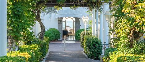 A pathway leading to a covered area with a piano, surrounded by lush greenery and white columns.