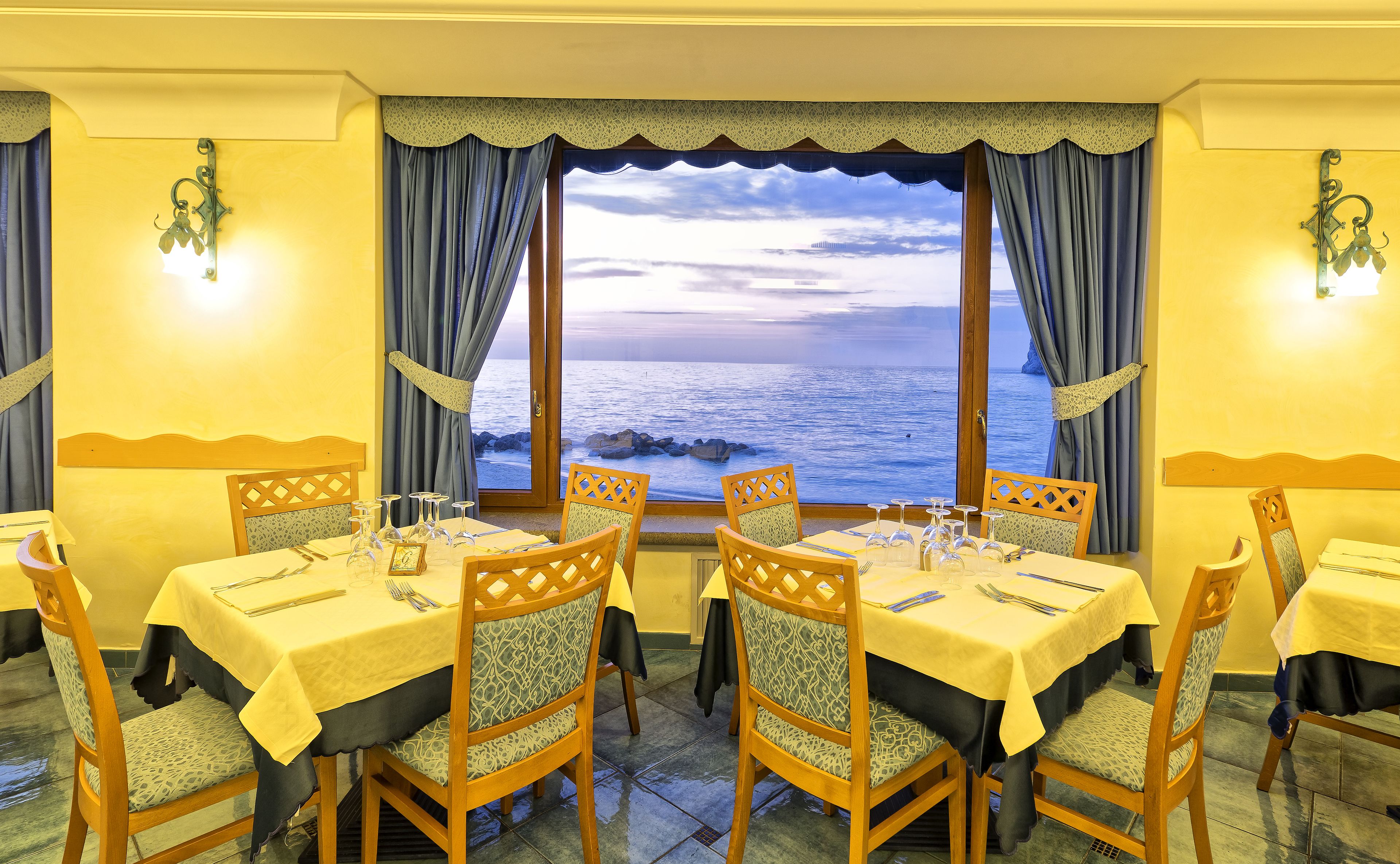 Un espace repas avec des tables dressées pour quatre personnes, des chaises dotées de coussins à motifs et une vue sur la mer grâce à de grandes fenêtres.