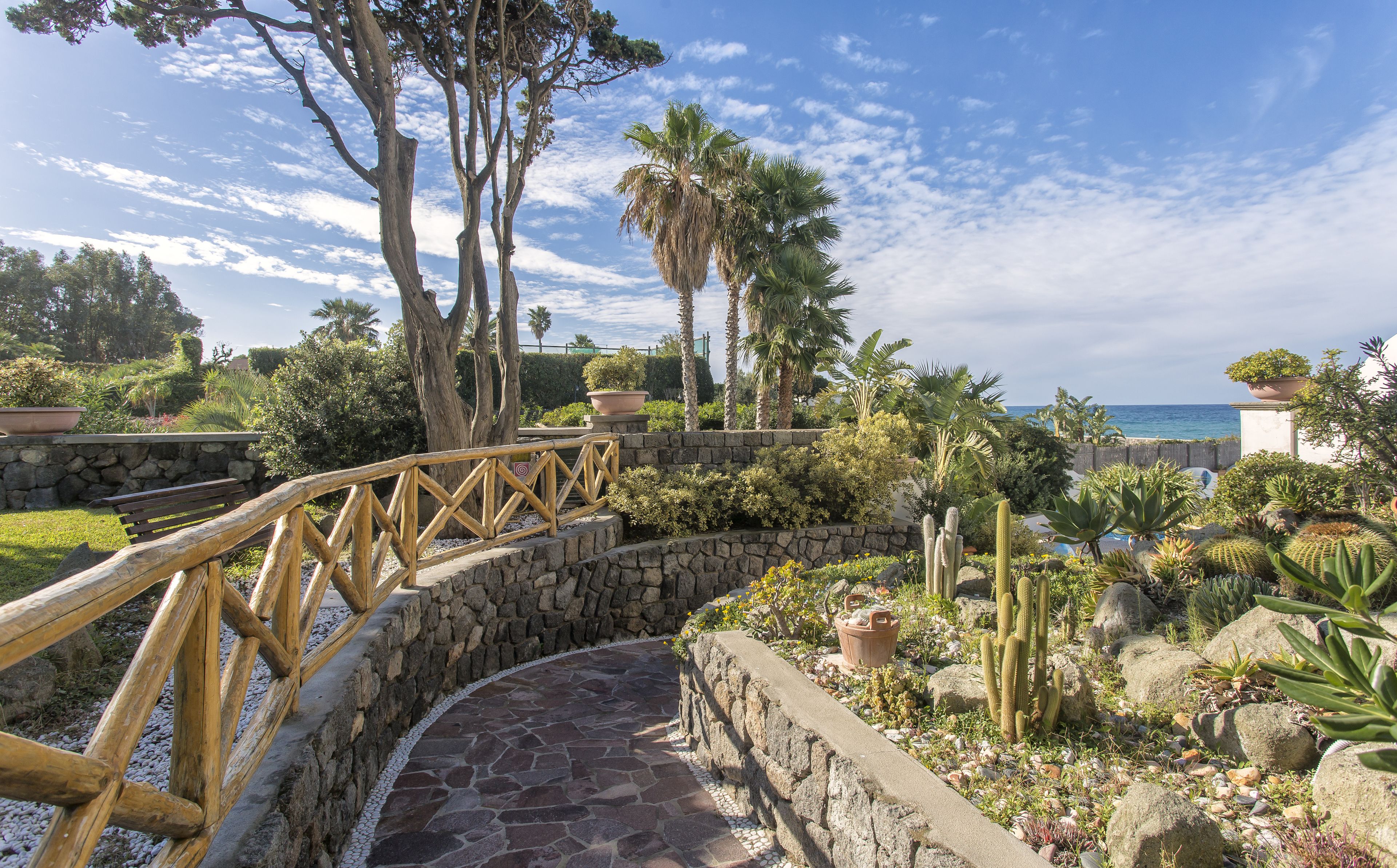 Un sentier de pierres mène à un pont en bois qui enjambe un jardin où poussent des cactus et des palmiers, offrant une vue sur une étendue d’eau.