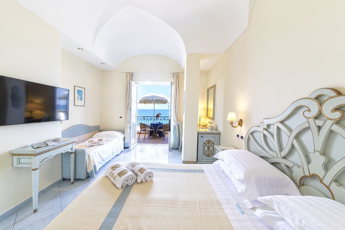 A hotel room with a bed, a desk, a television, and a view of the sea through an open door.