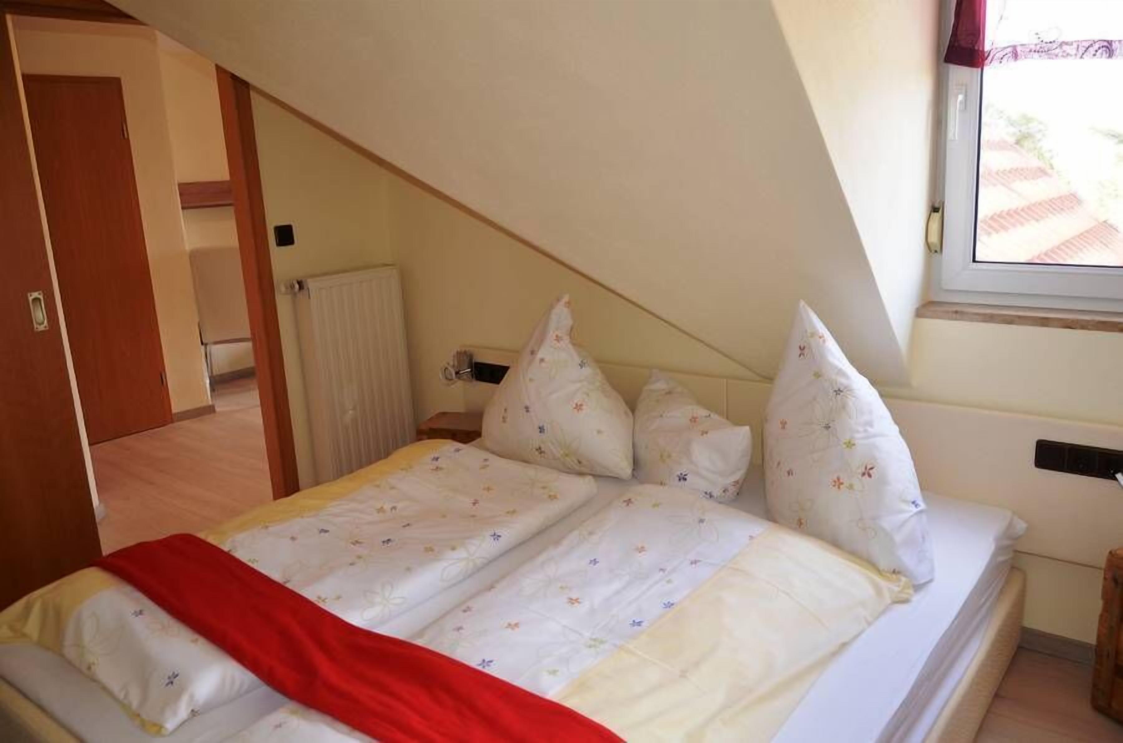 Un dormitorio con techo inclinado, cama con ropa de cama floral, manta roja y ventana con vistas a edificios.