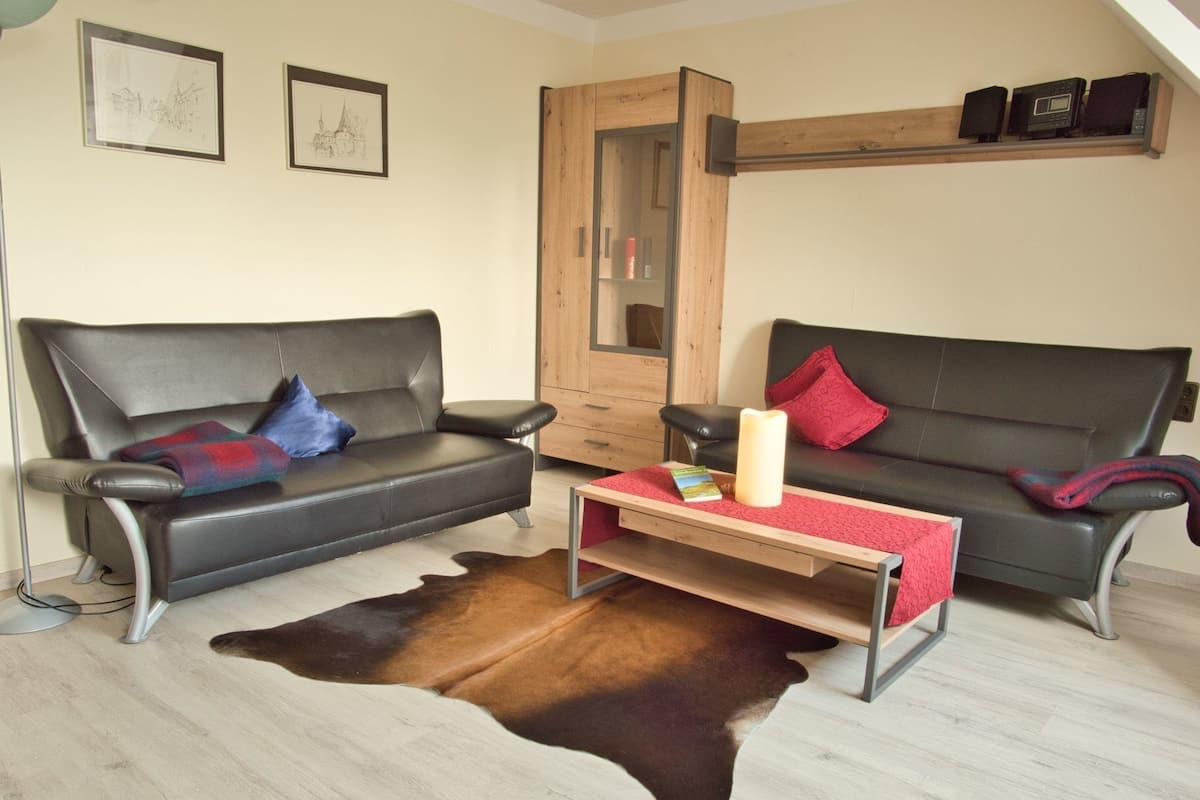 A living room with two black sofas, a wooden cabinet, a glass door, and a small table with a book and candle.