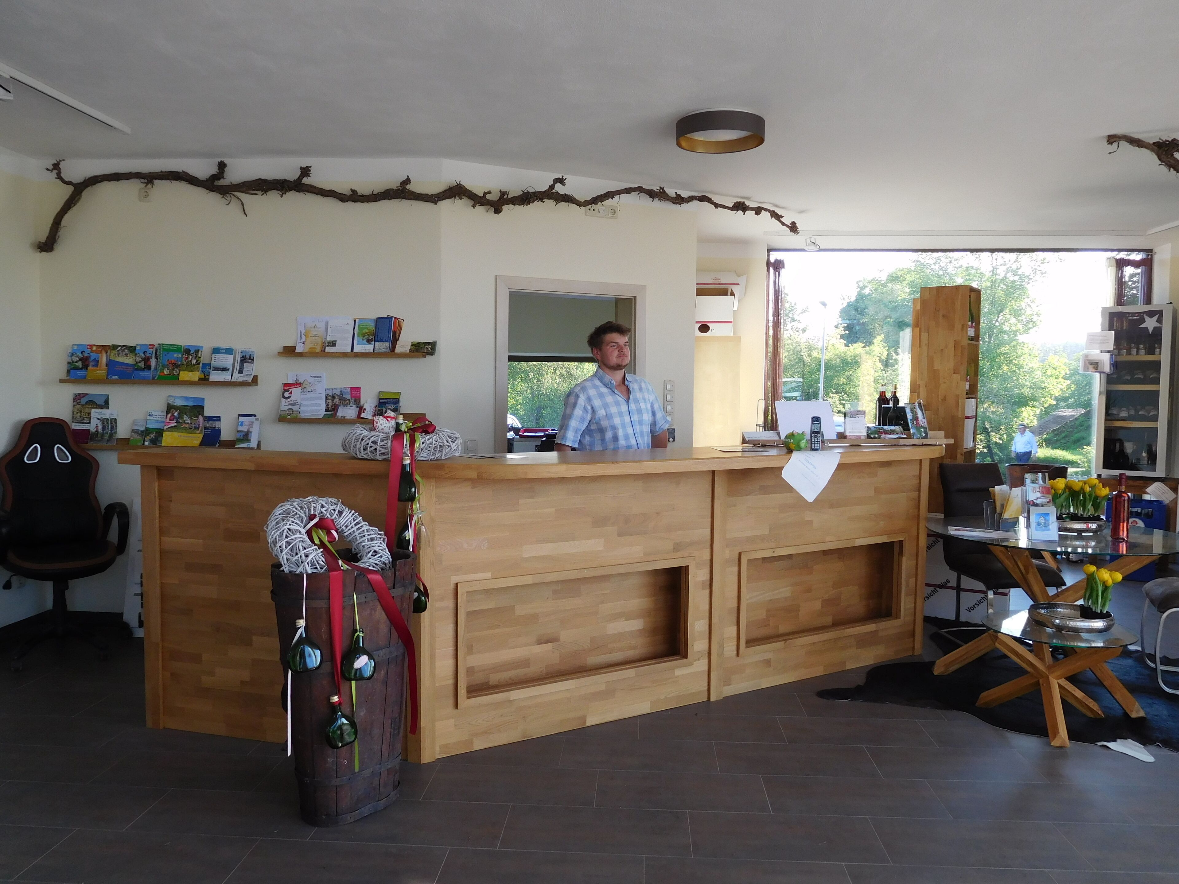 A reception desk with a wooden counter, a man standing behind it, and a view of the outdoors through a window.