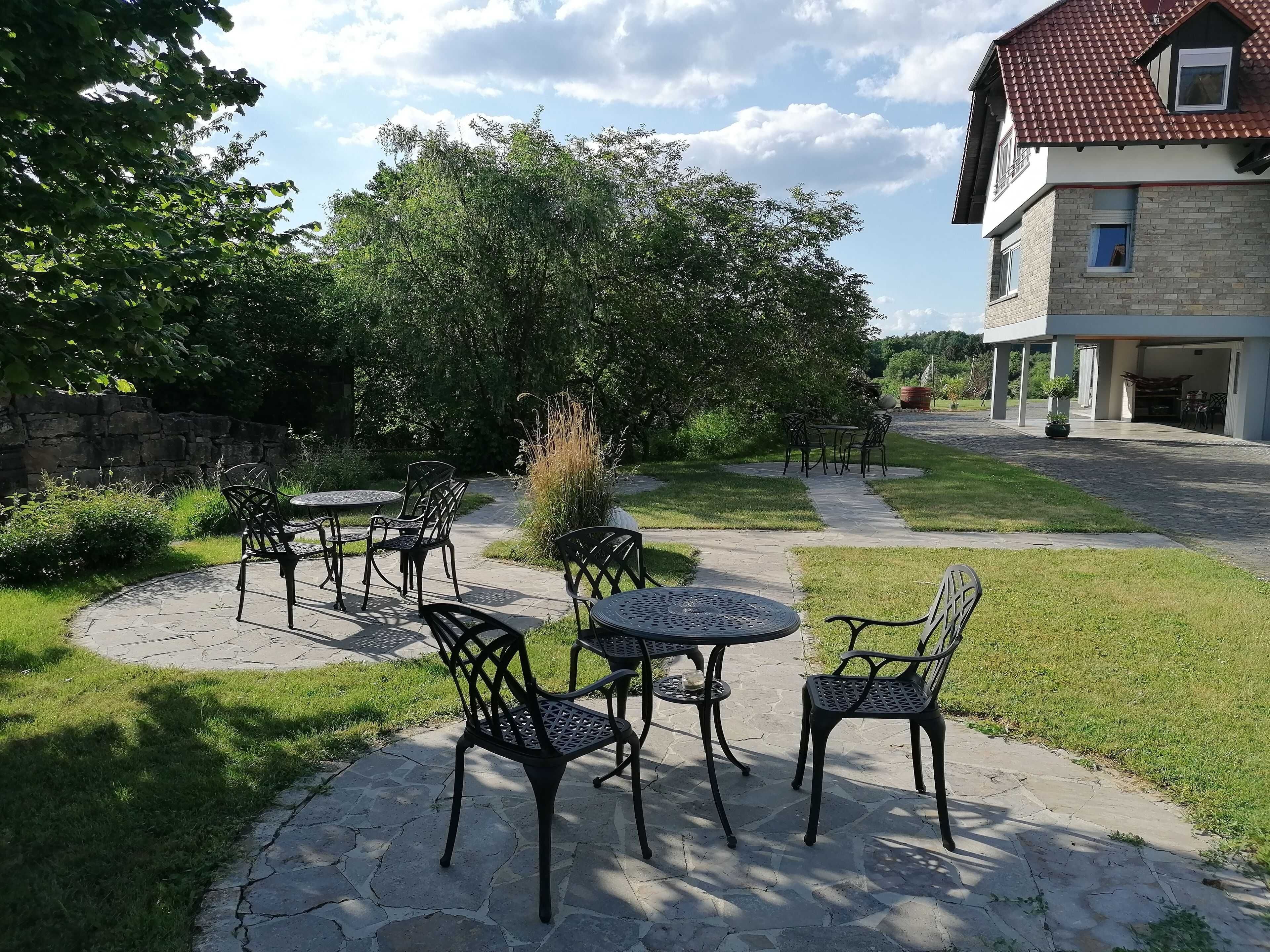 Area patio esterna con pavimento in pietra, sedie e tavoli in metallo, circondata da vegetazione e un muro di pietra.