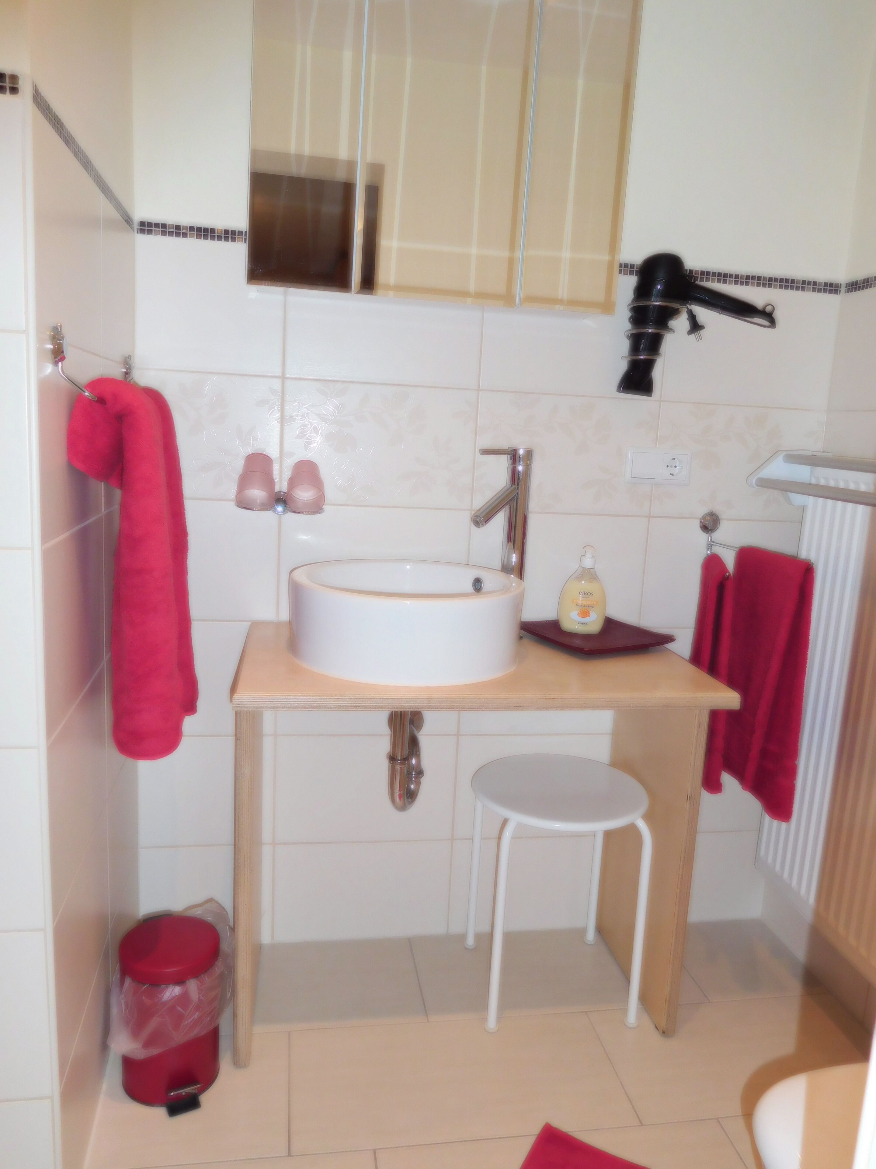 Casa de banho com uma pia branca, espelho, toalha vermelha, secador de cabelo e um lixo vermelho.