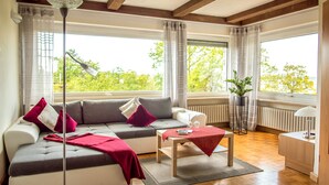 A modern living room with a sofa, a coffee table, and a floor lamp. There are large windows with curtains, a radiator, and a potted plant. - Kirschberghof - Adults Only (Sommerhausen)