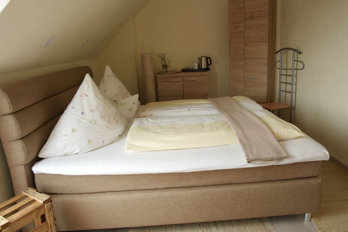 A bedroom with a single bed, a wooden wardrobe, a small table with a kettle, and a ladder against the wall.