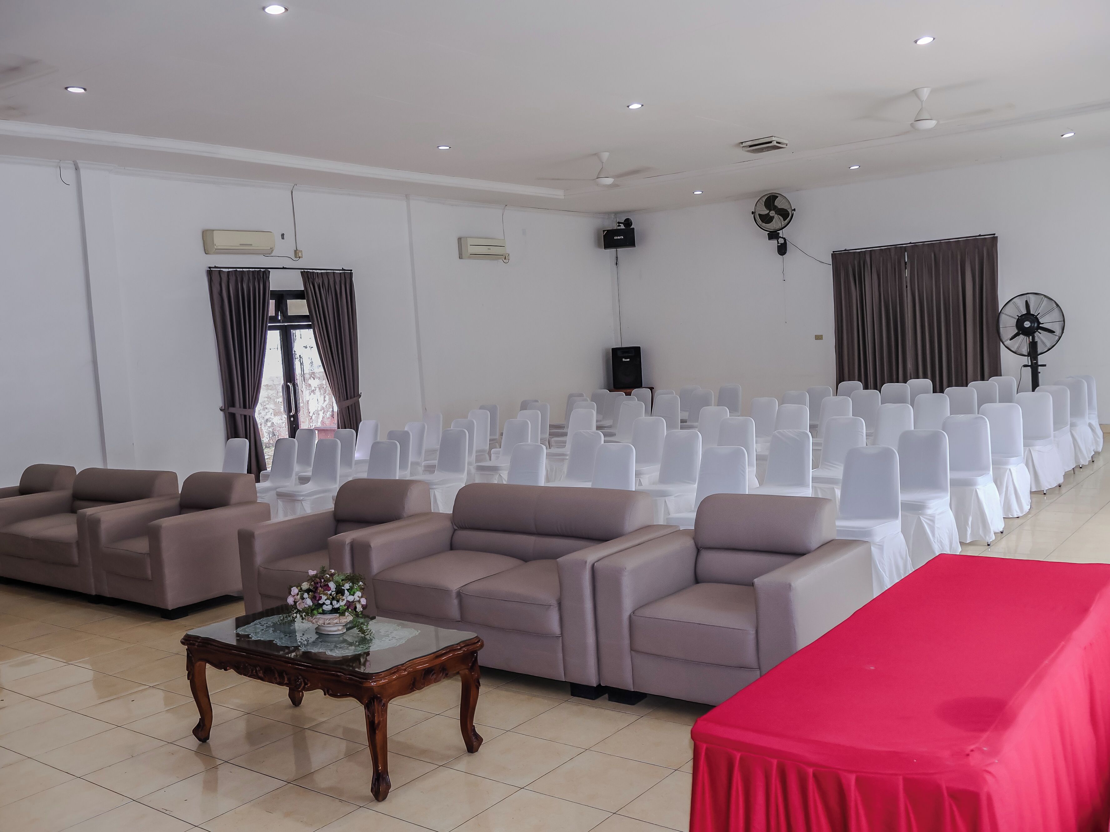 Una sala conferenze con file di sedie bianche, un palco con un tavolo rosso e una ventola a parete.