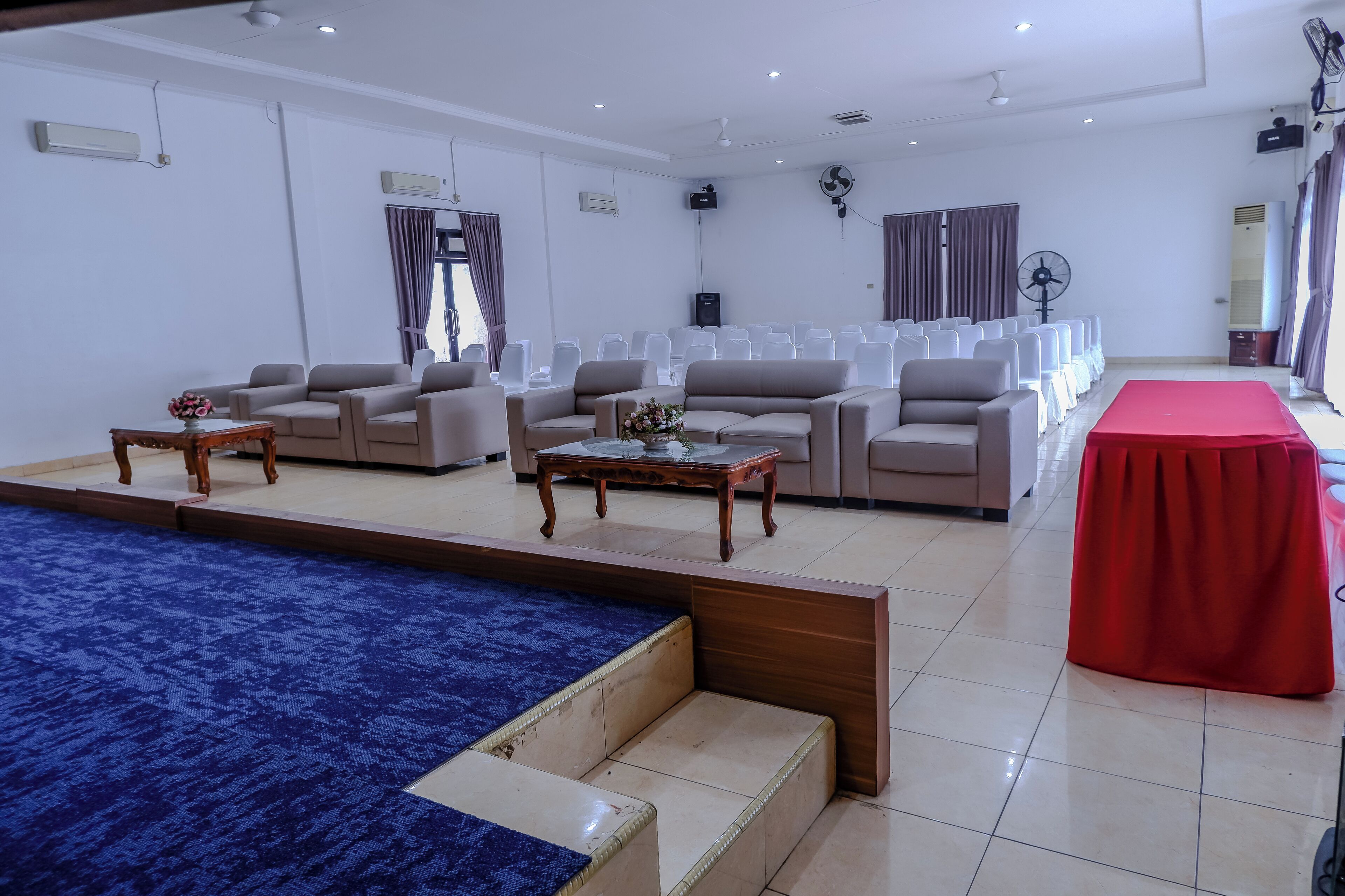 A spacious room with a blue carpet, beige sofas, a red table, and a fan.