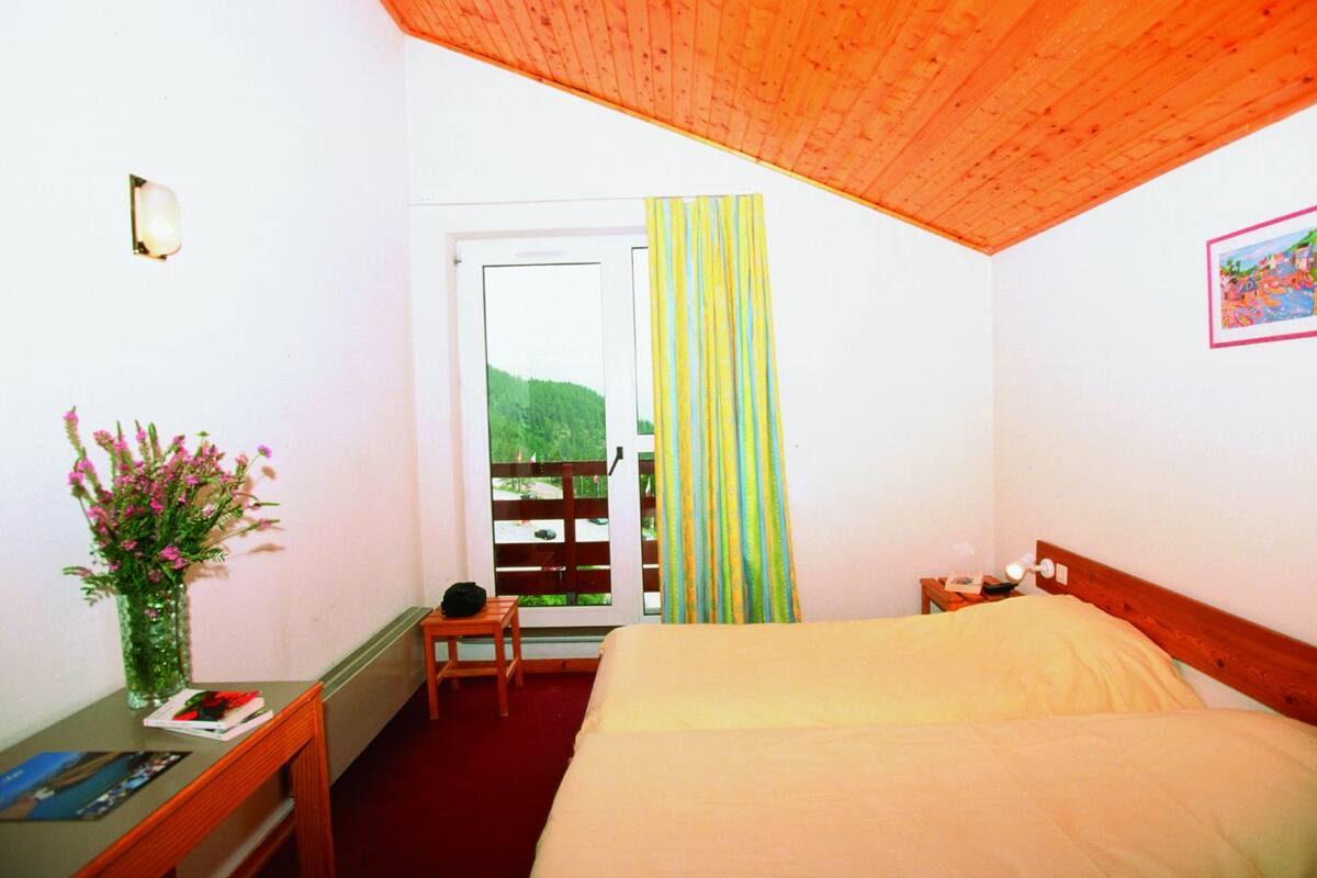 A bedroom with a wooden ceiling, a bed, a nightstand, a small table with a vase of flowers, a chair, and a window with a view of greenery.