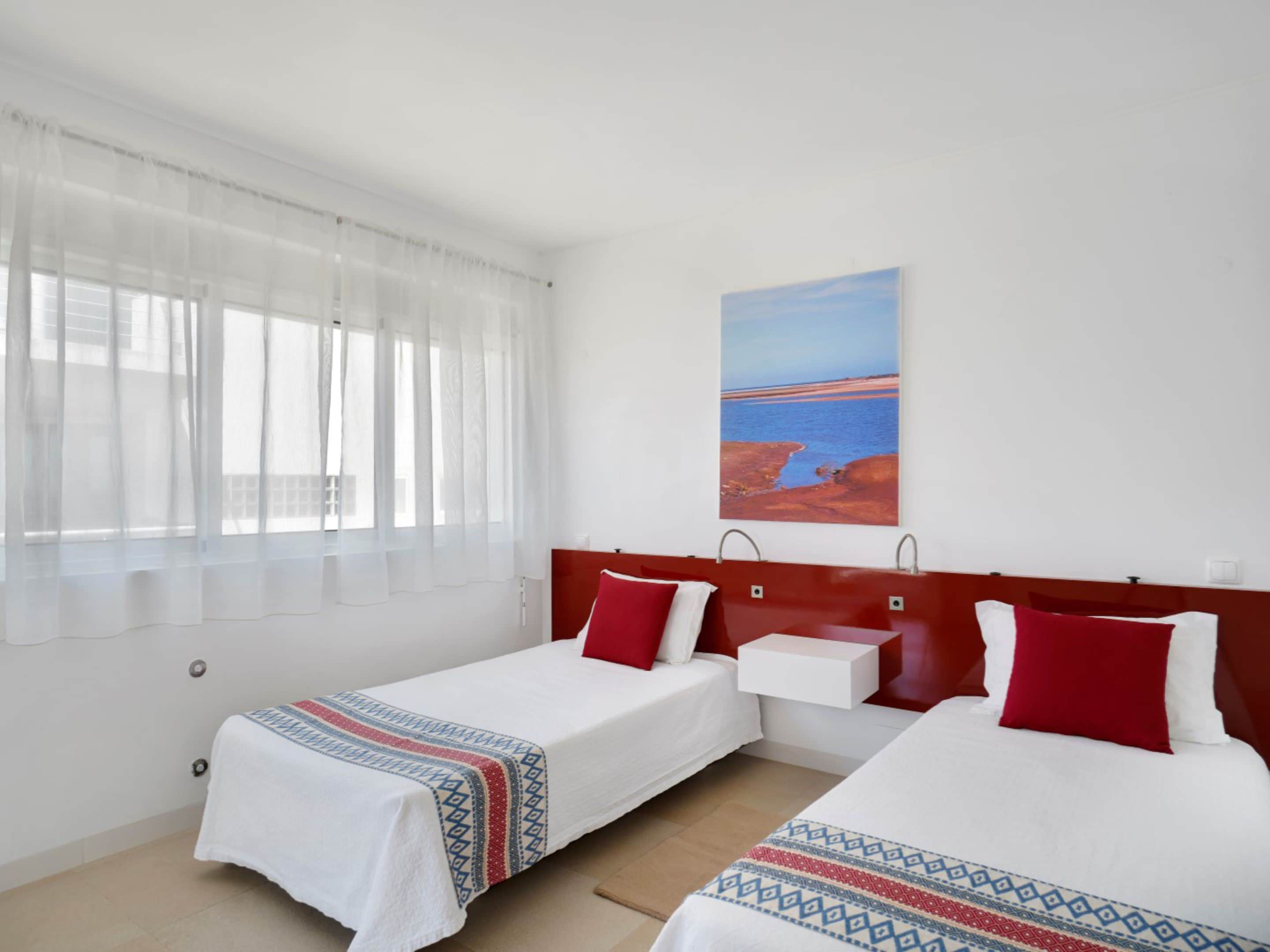 A hotel room with two single beds, a window with sheer curtains, a painting of a coastal landscape, and a red headboard.