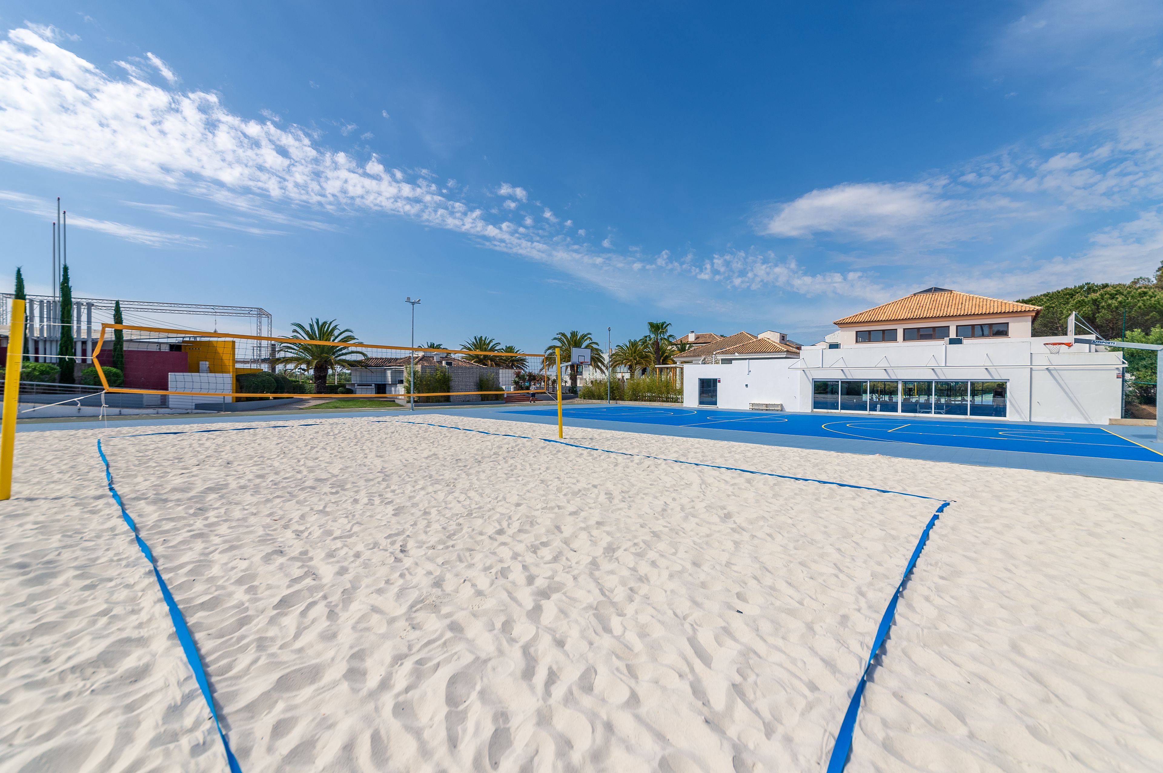Un terrain de beach-volley sablonneux, doté d’une surface bleue, entouré d’immeubles et de palmiers.
