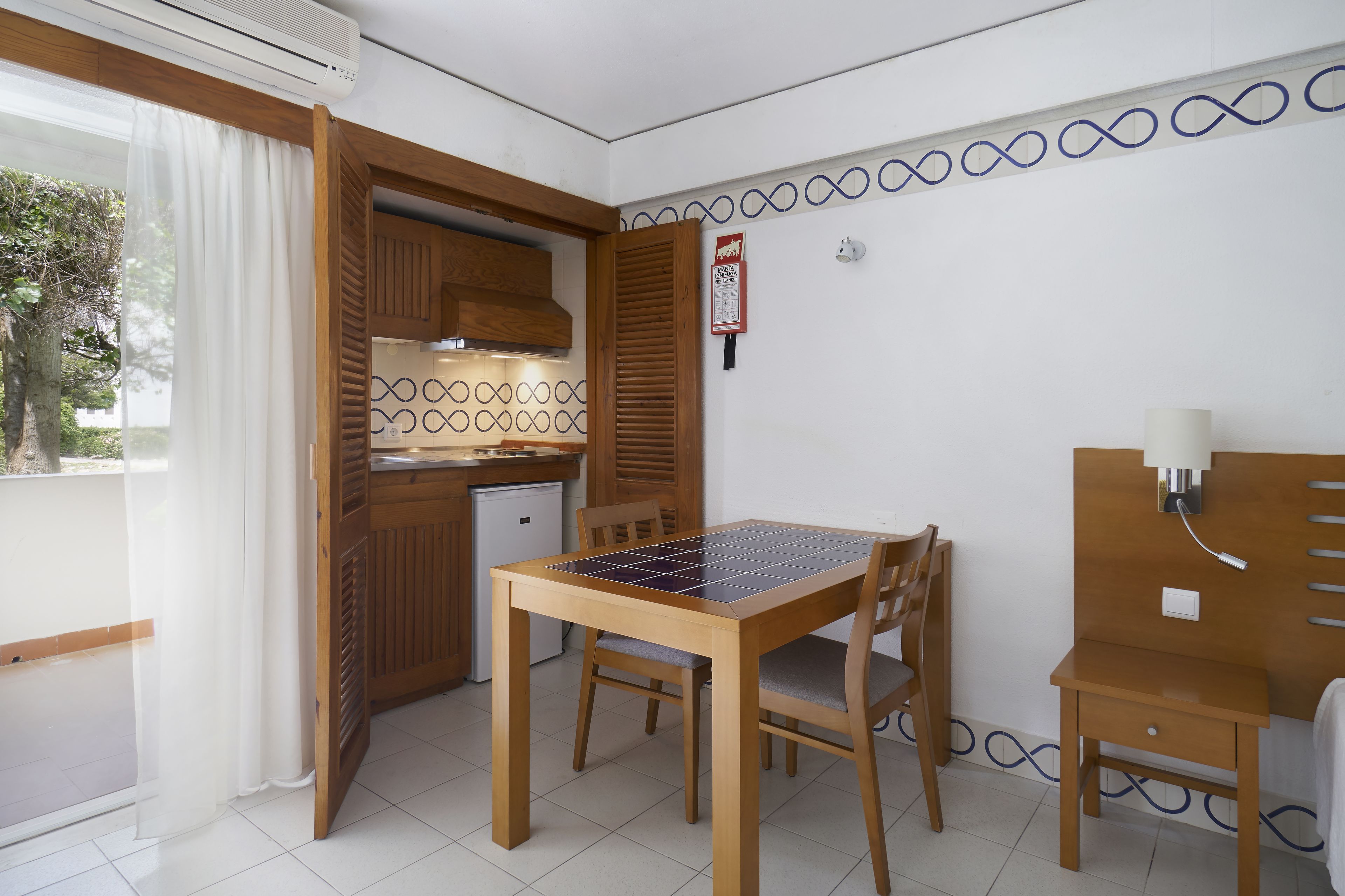 A compact room with a dining table, chairs, a small kitchenette, and a window with curtains.