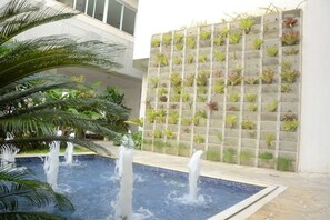 A pool with water jets, surrounded by palm trees and a wall with potted plants. - Charmoso Confortável Flat (Cabo Frio)