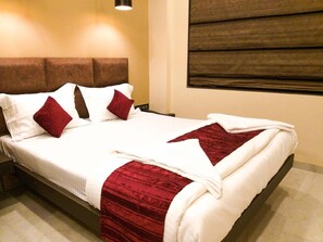 A neatly made bed with white linens and red accents, a brown headboard, and a window with blinds.