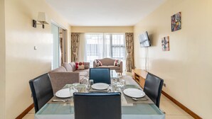 A dining area with a glass table set for four, a sofa, and a TV mounted on the wall. - Lenana Suites (Nairobi)