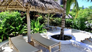 A tropical outdoor lounge area with wooden loungers, a thatched roof structure, and a central fire pit surrounded by plants. - Nyumbani Residence Apartments (Jambiani)