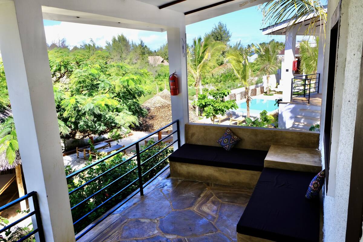 A balcony with a stone floor, a black cushioned bench, and a view of a pool and lush greenery.