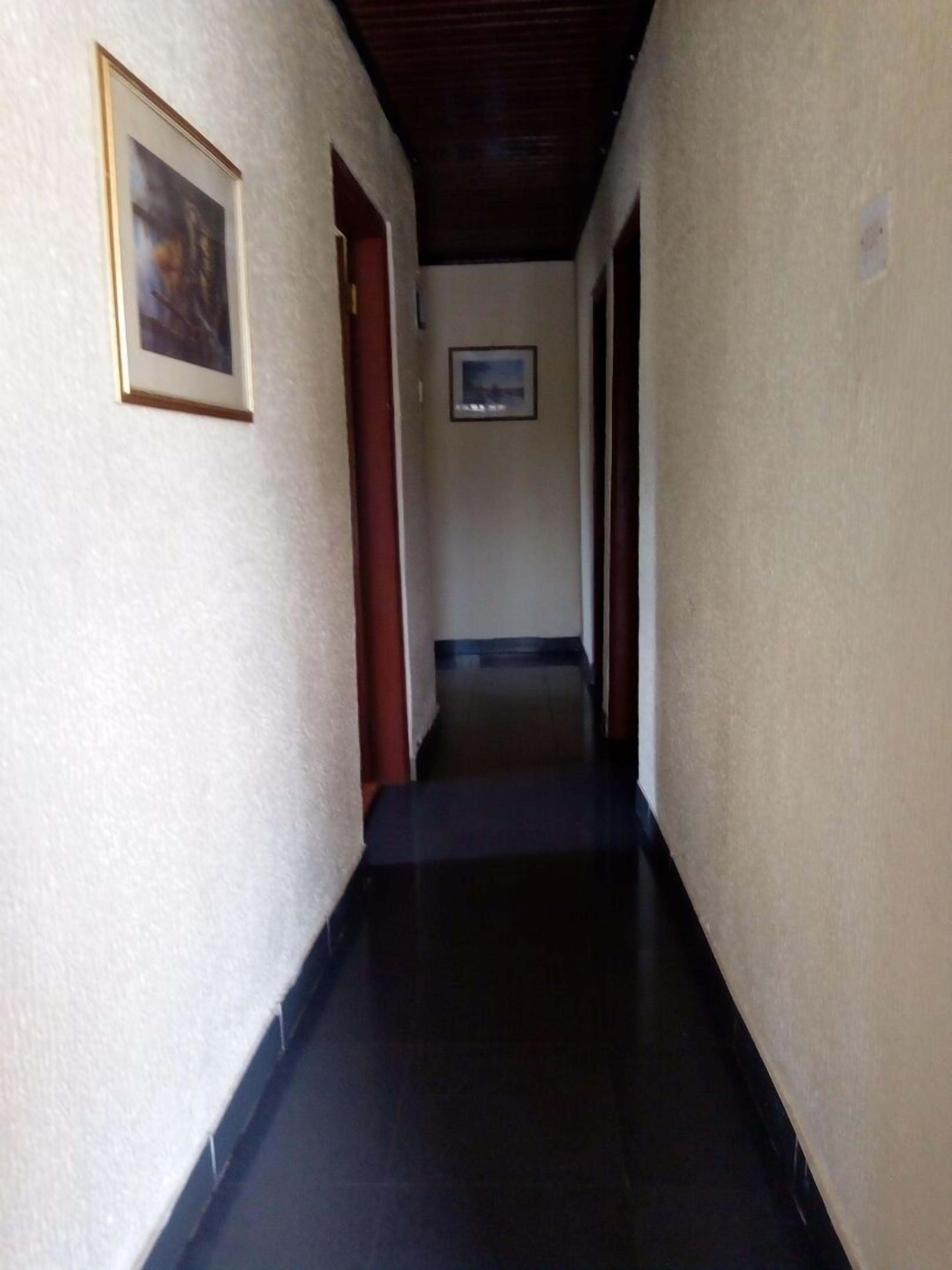 A narrow hallway with a dark floor, light walls, and a wooden ceiling. There are framed pictures on the walls and a door at the end.