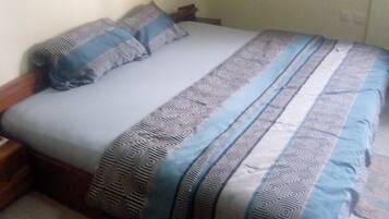 Una cama con colcha y almohadas estampadas, una planta colgada en la pared y un suelo de baldosas.