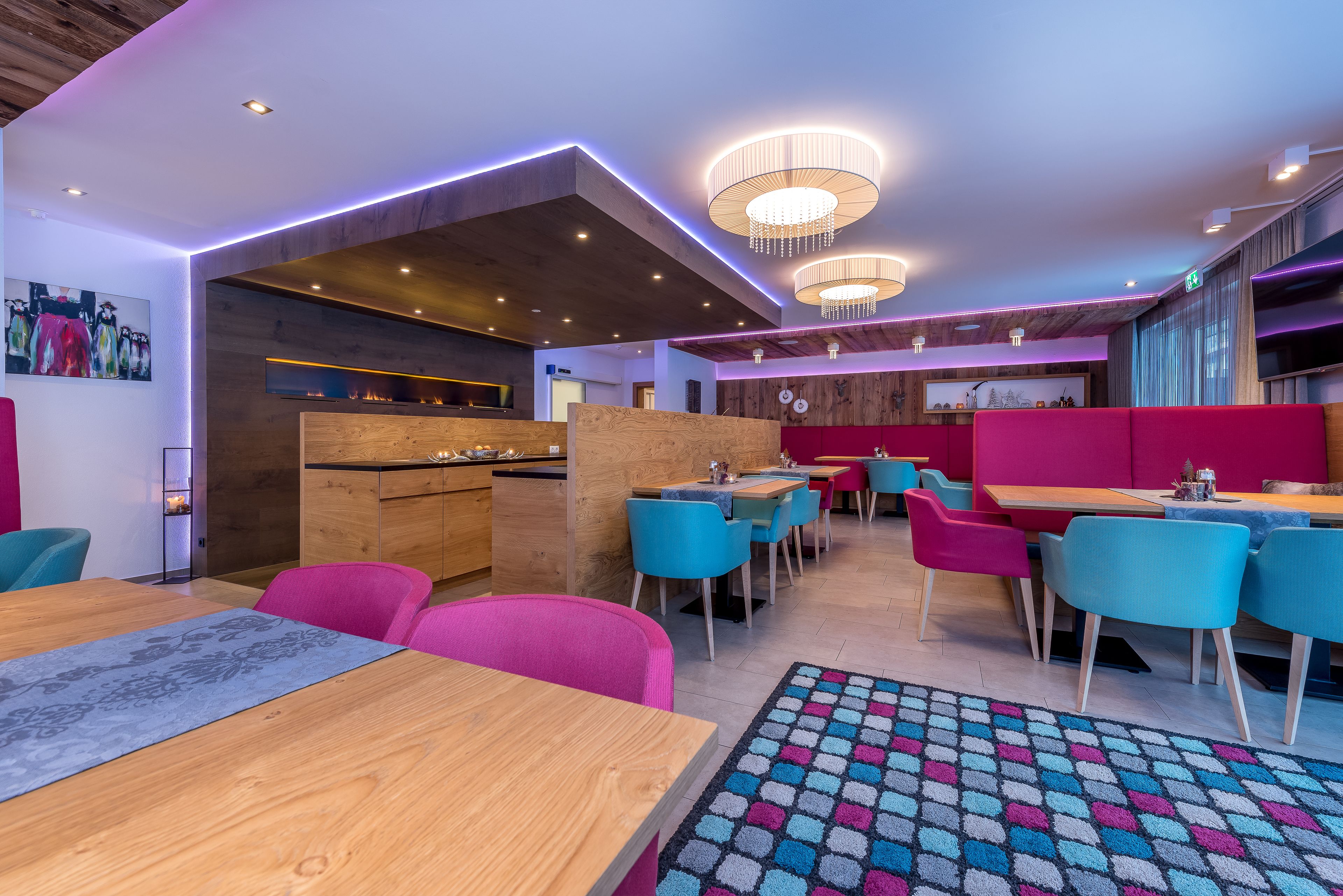 A modern restaurant with wooden and colorful furniture, pendant lighting, and a patterned rug.