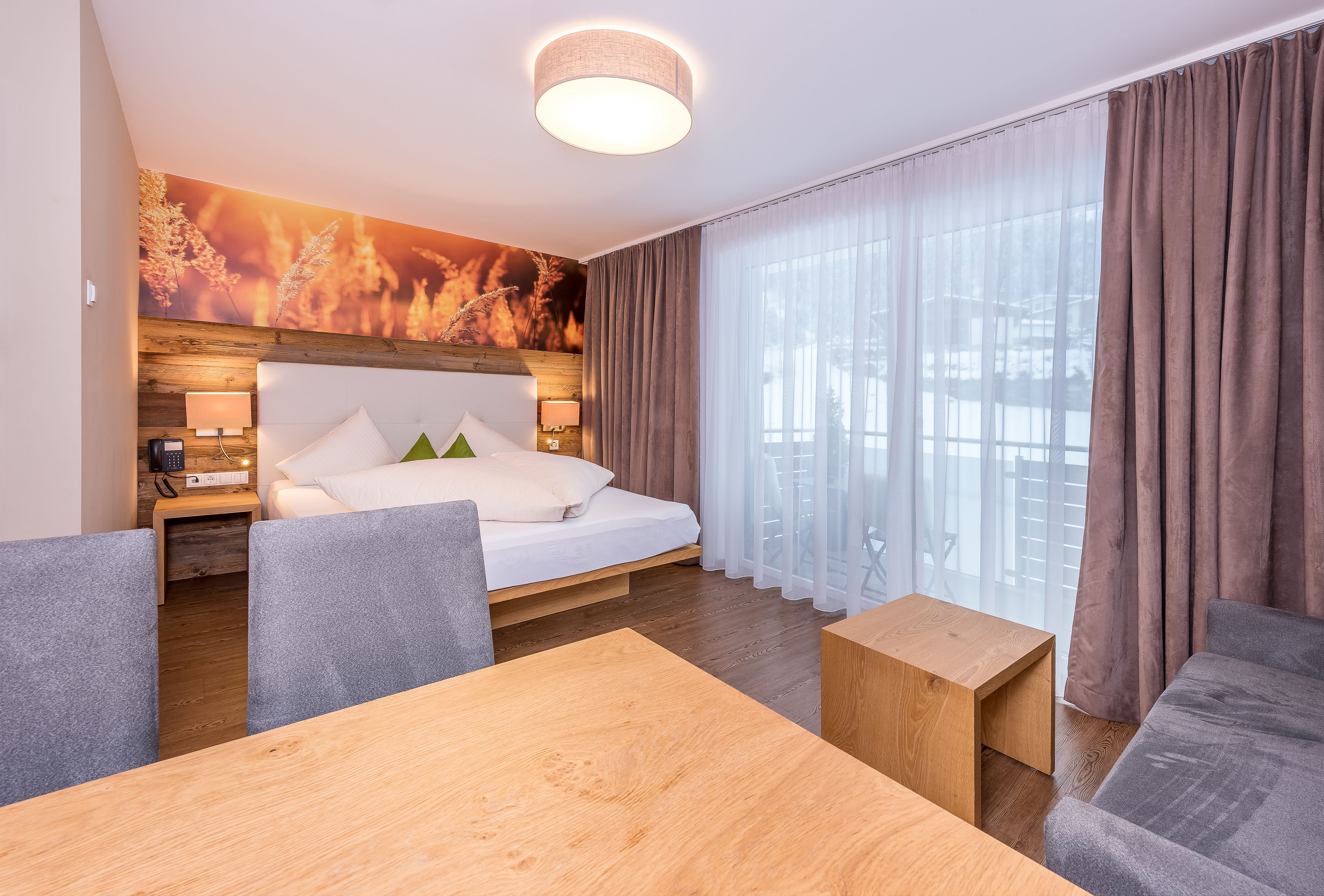 A modern hotel room with a wooden headboard, a large bed, a dining table, and a sofa.