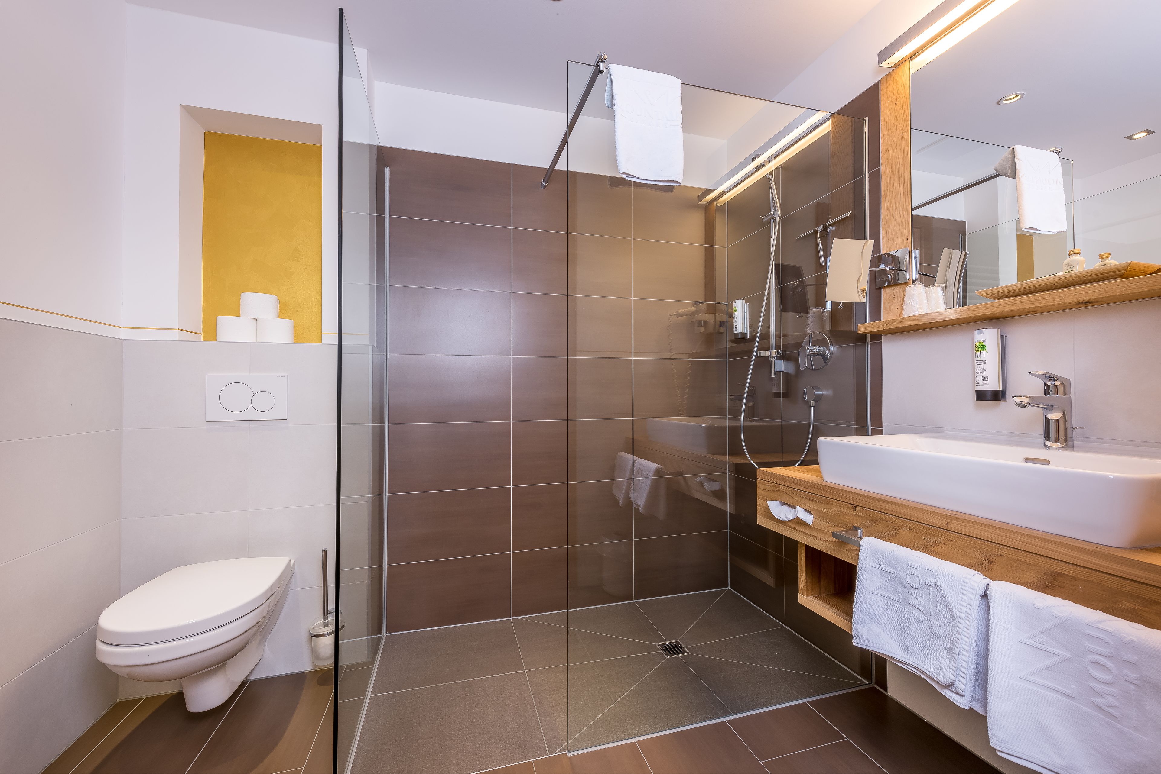 Un baño moderno con ducha envuelta en vidrio, inodoro blanco, tocador de madera con lavamanos y un espejo grande.