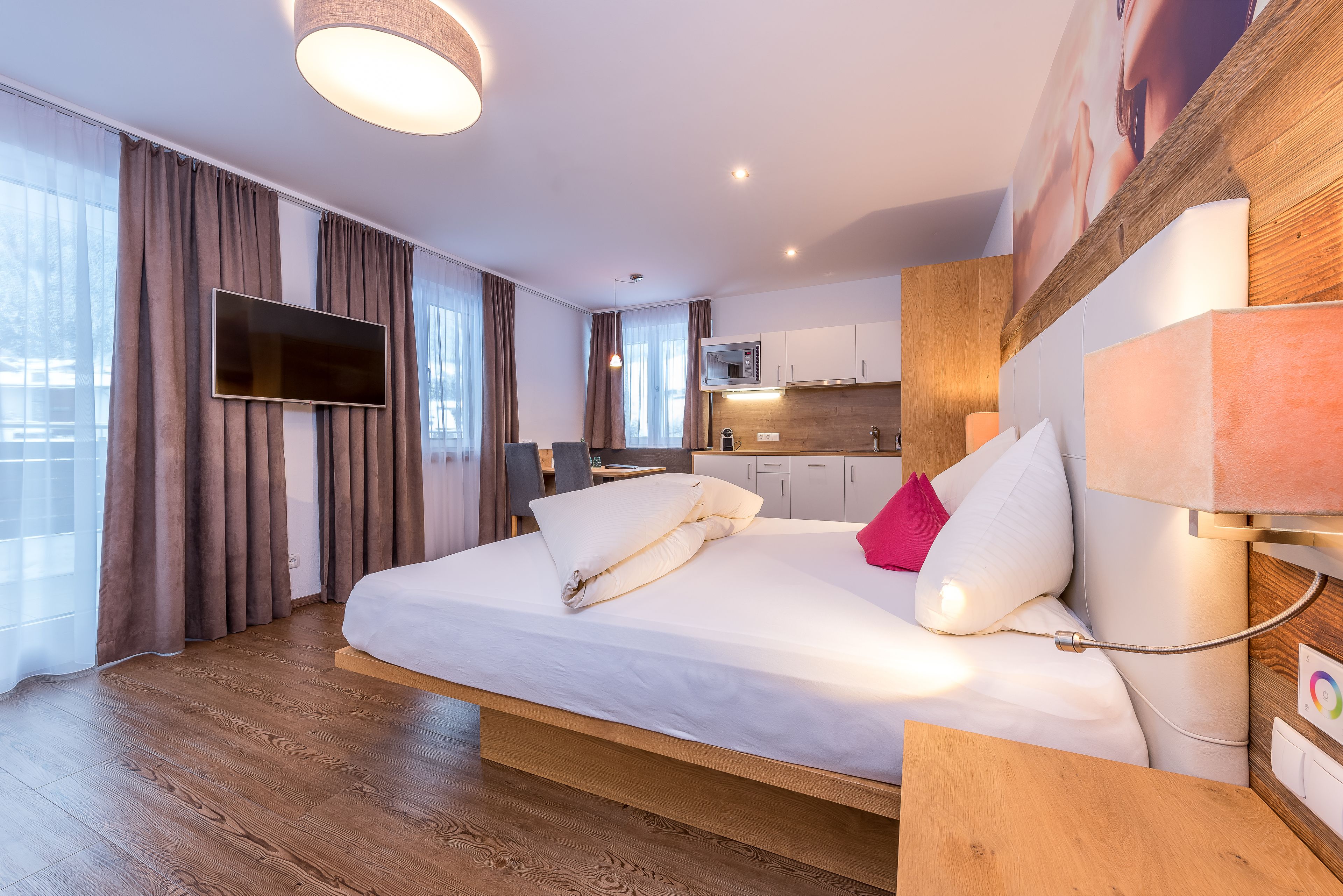 A modern hotel room with a large bed, a flat-screen TV, a wooden floor, a kitchen area, and a large window with curtains.