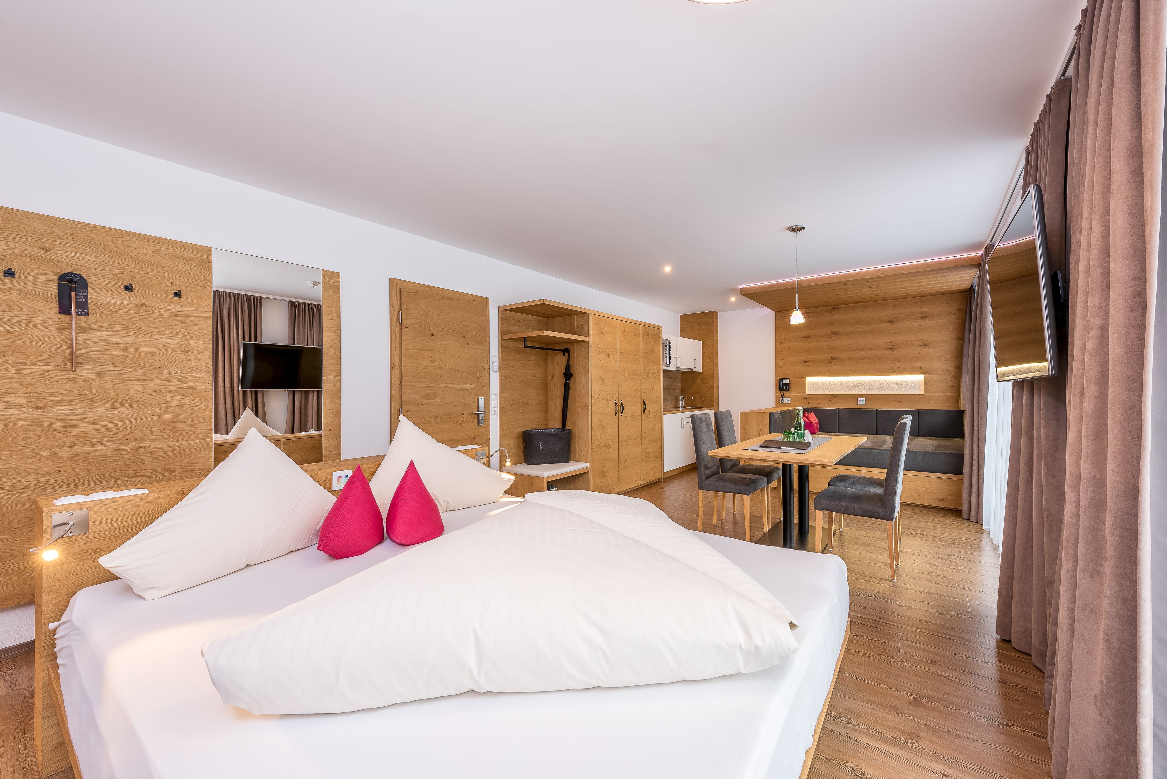 A modern hotel room with a large bed, wooden walls, a dining area, and a TV.