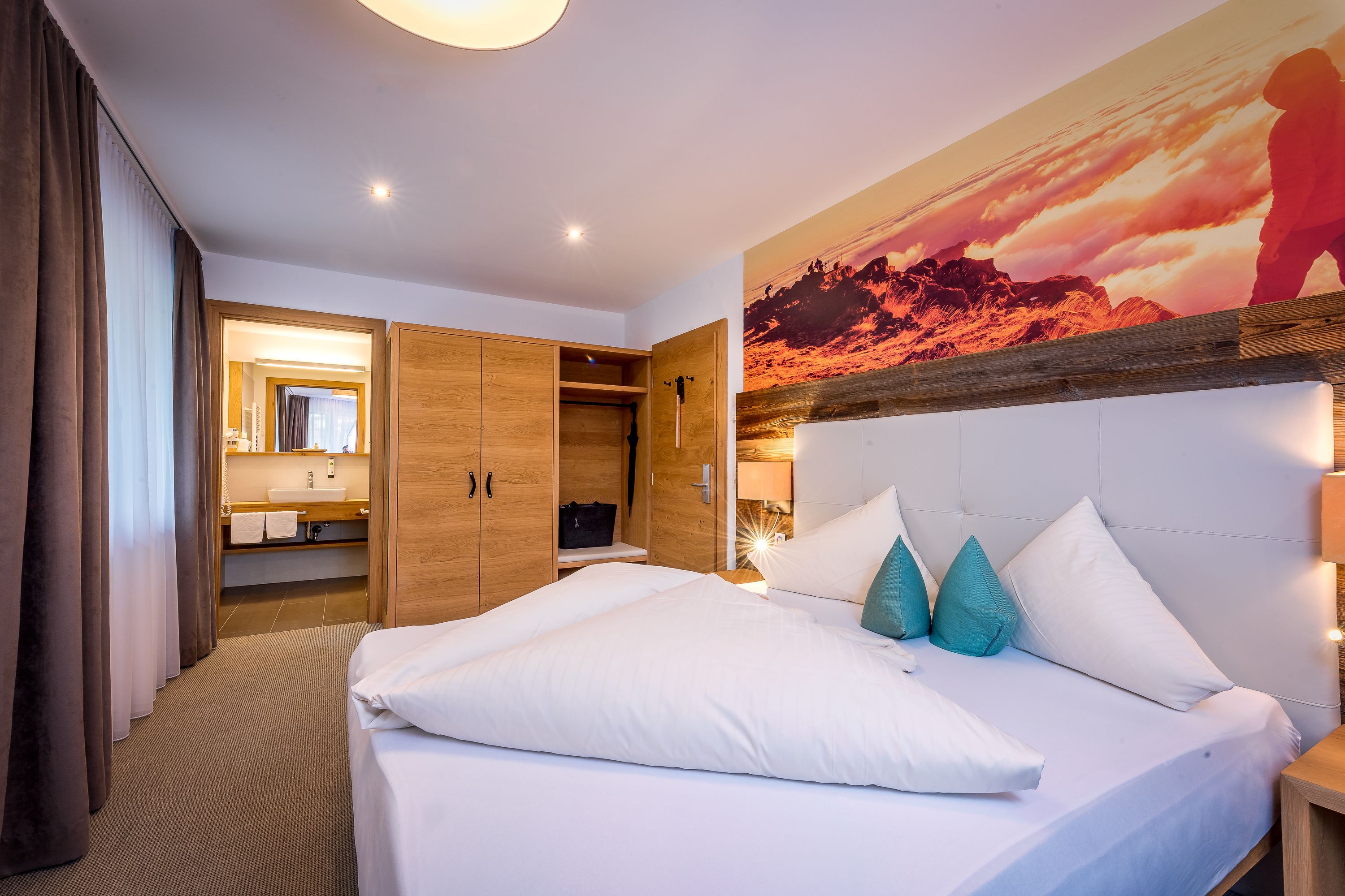 A hotel room with a large bed, a wooden wardrobe, a bathroom with a mirror and sink, and a sunset mural on the wall.