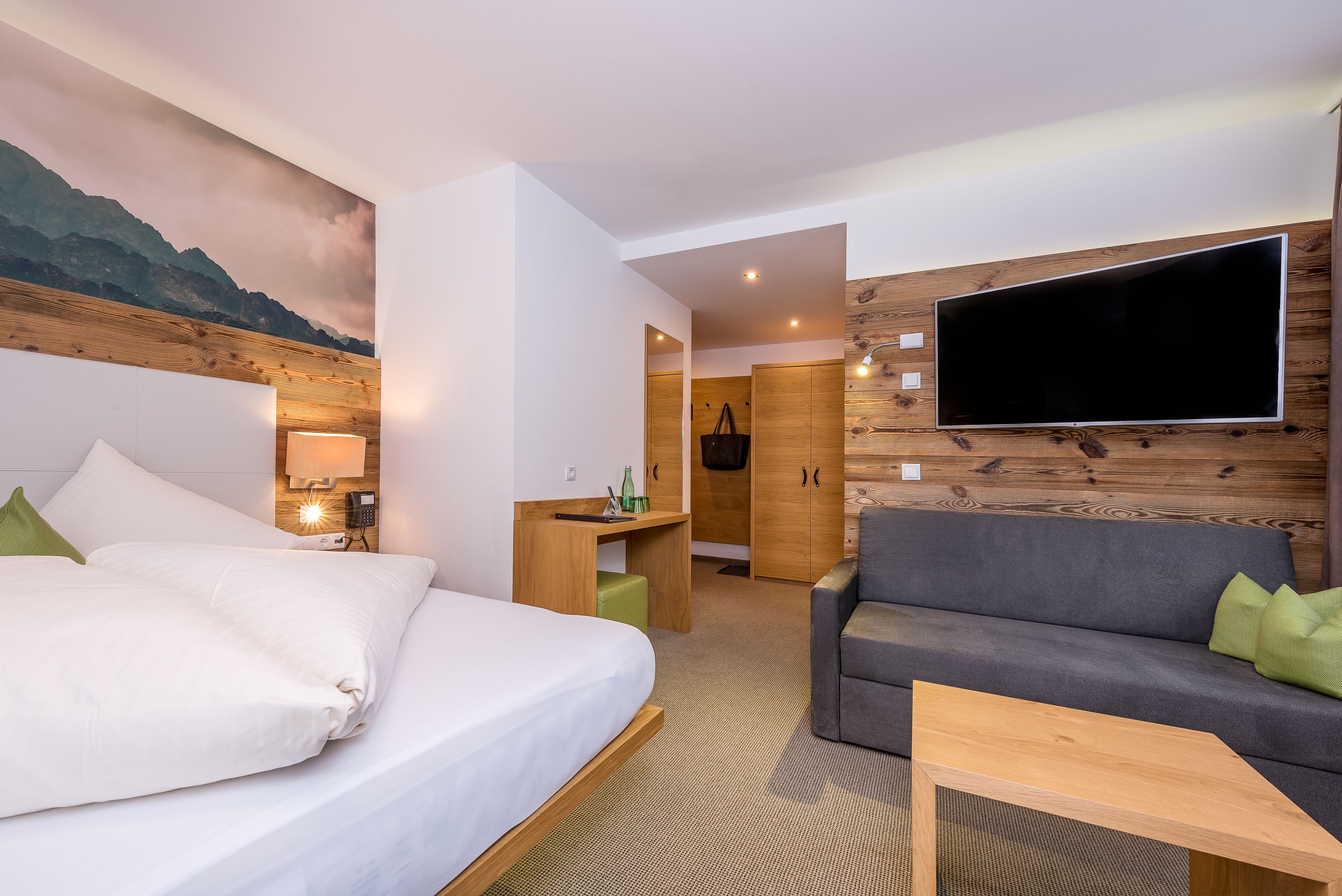 A hotel room with a bed, a sofa, a TV mounted on the wall, a desk, and a wooden panel design.