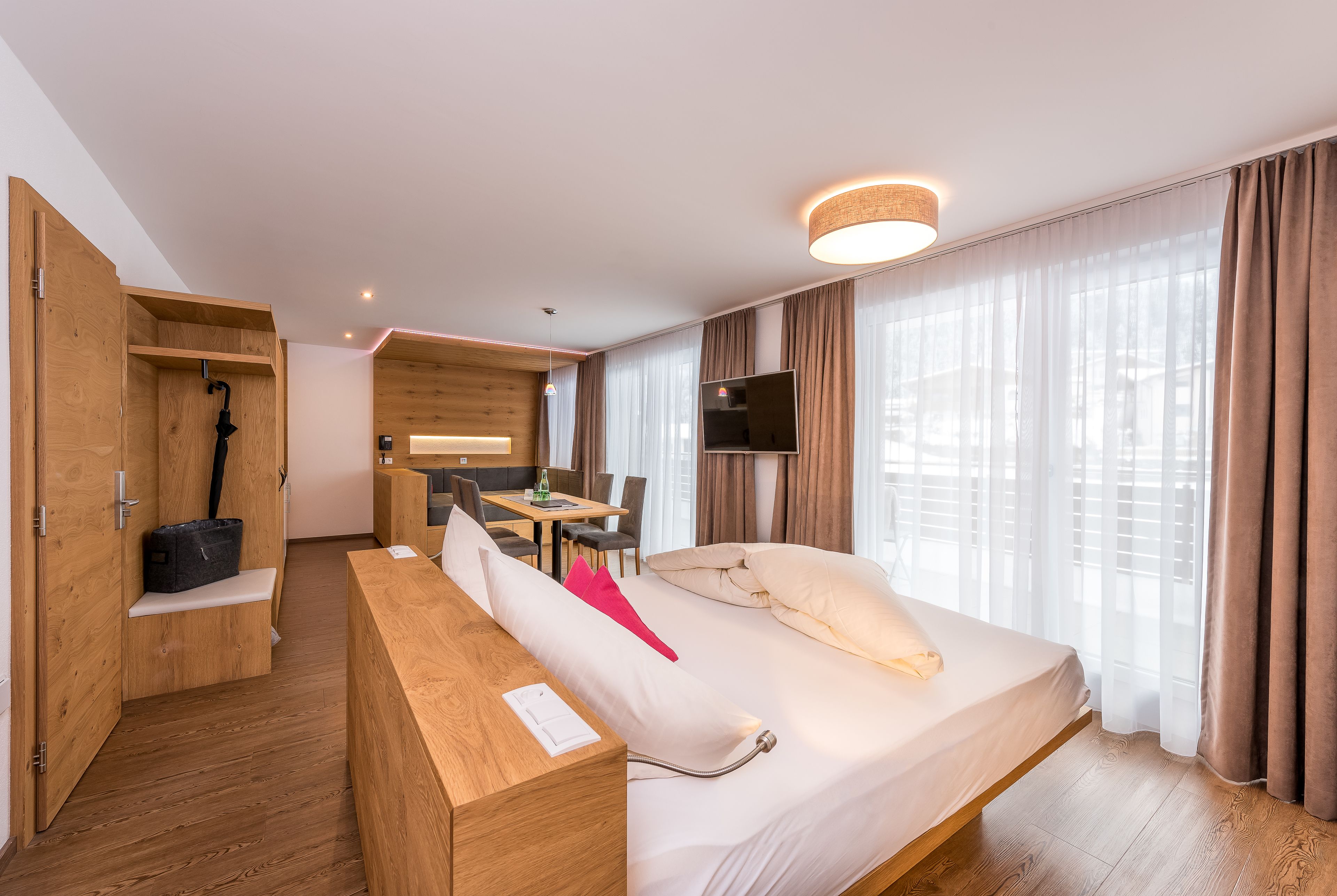 A modern hotel room with a wooden floor, a bed with white bedding, a desk with a chair, a television, and a large window with curtains.