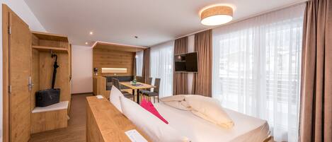 A modern hotel room with a wooden floor, a bed with white bedding, a desk with a chair, a television, and a large window with curtains.