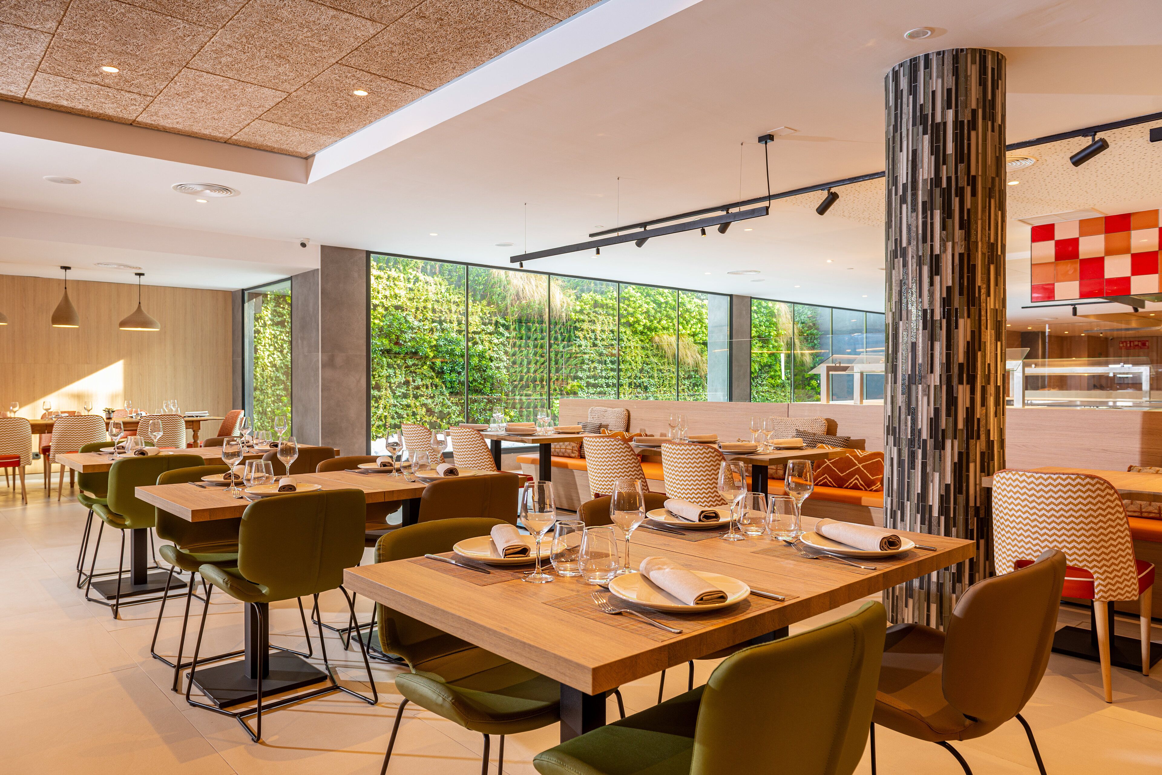 Un comedor moderno con mesas de madera y sillas verdes, ventanas amplias que ofrecen vistas a la vegetación, y un pilar decorado con un diseño llamativo.