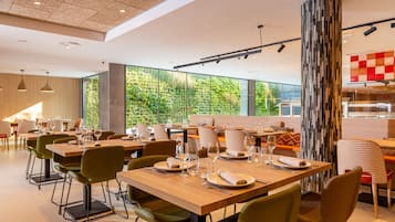 Un comedor moderno con mesas de madera y sillas verdes, ventanas amplias que ofrecen vistas a la vegetación, y un pilar decorado con un diseño llamativo.