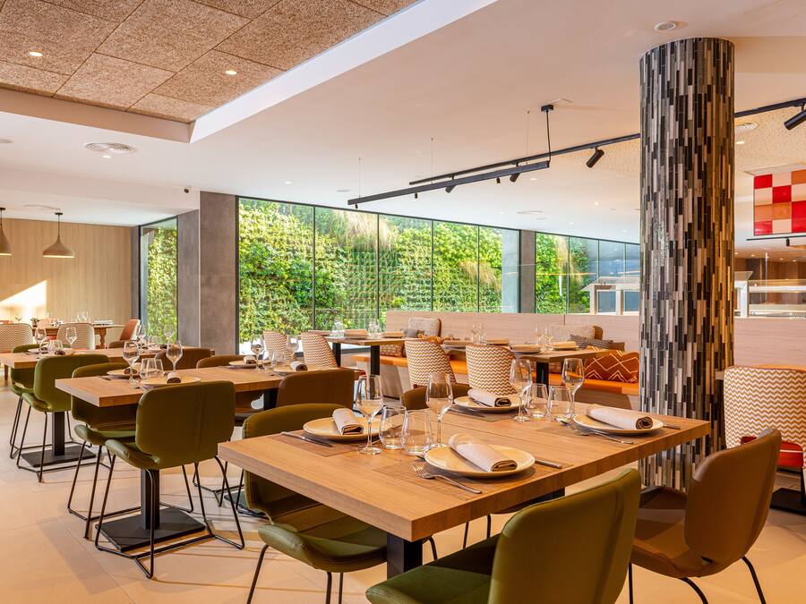 Un comedor moderno con mesas de madera y sillas verdes, ventanas amplias que ofrecen vistas a la vegetación, y un pilar decorado con un diseño llamativo.