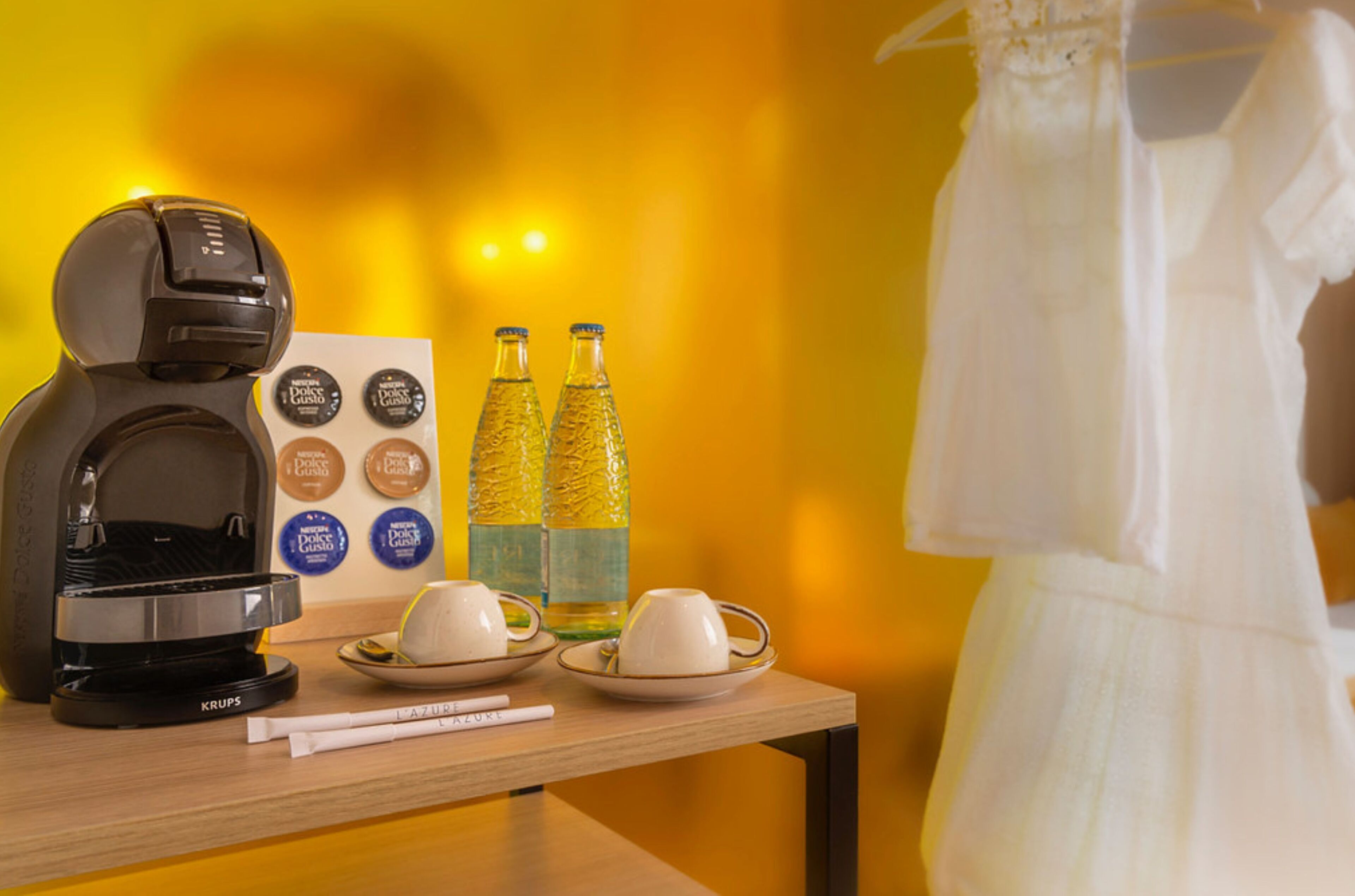 Une machine à café, deux bouteilles d’eau et une robe sont posés sur une table, avec un mur jaune en arrière-plan.