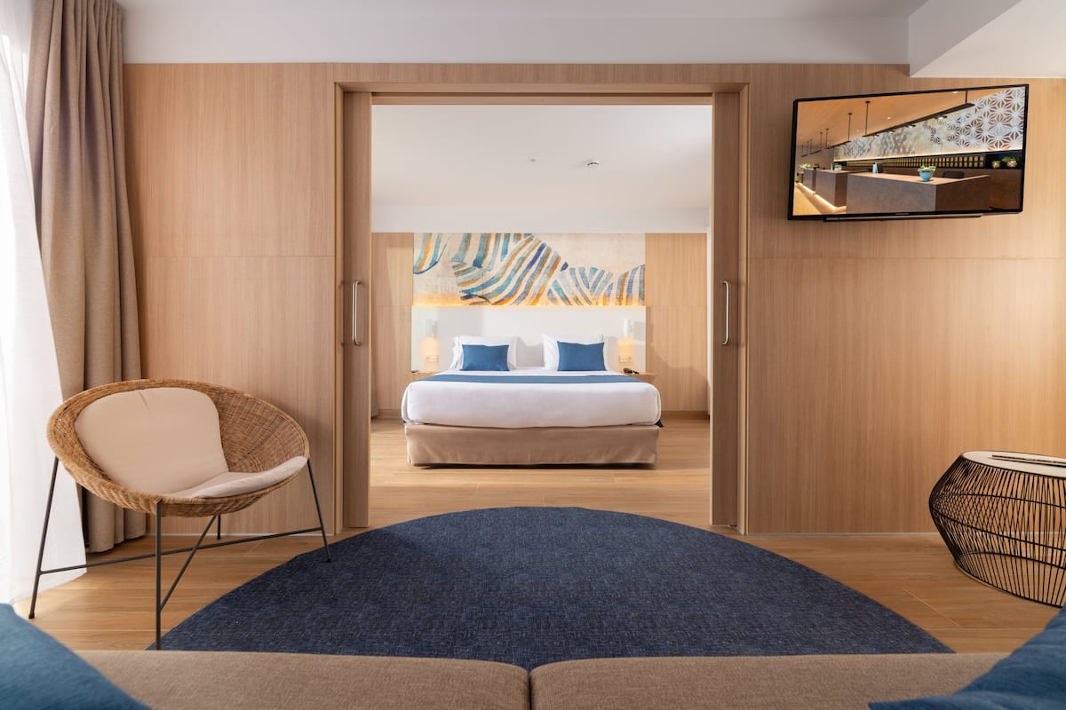 Une chambre d’hôtel moderne avec un grand lit, un fauteuil en osier, un téléviseur à écran plat et une vue sur le paysage urbain par la fenêtre.